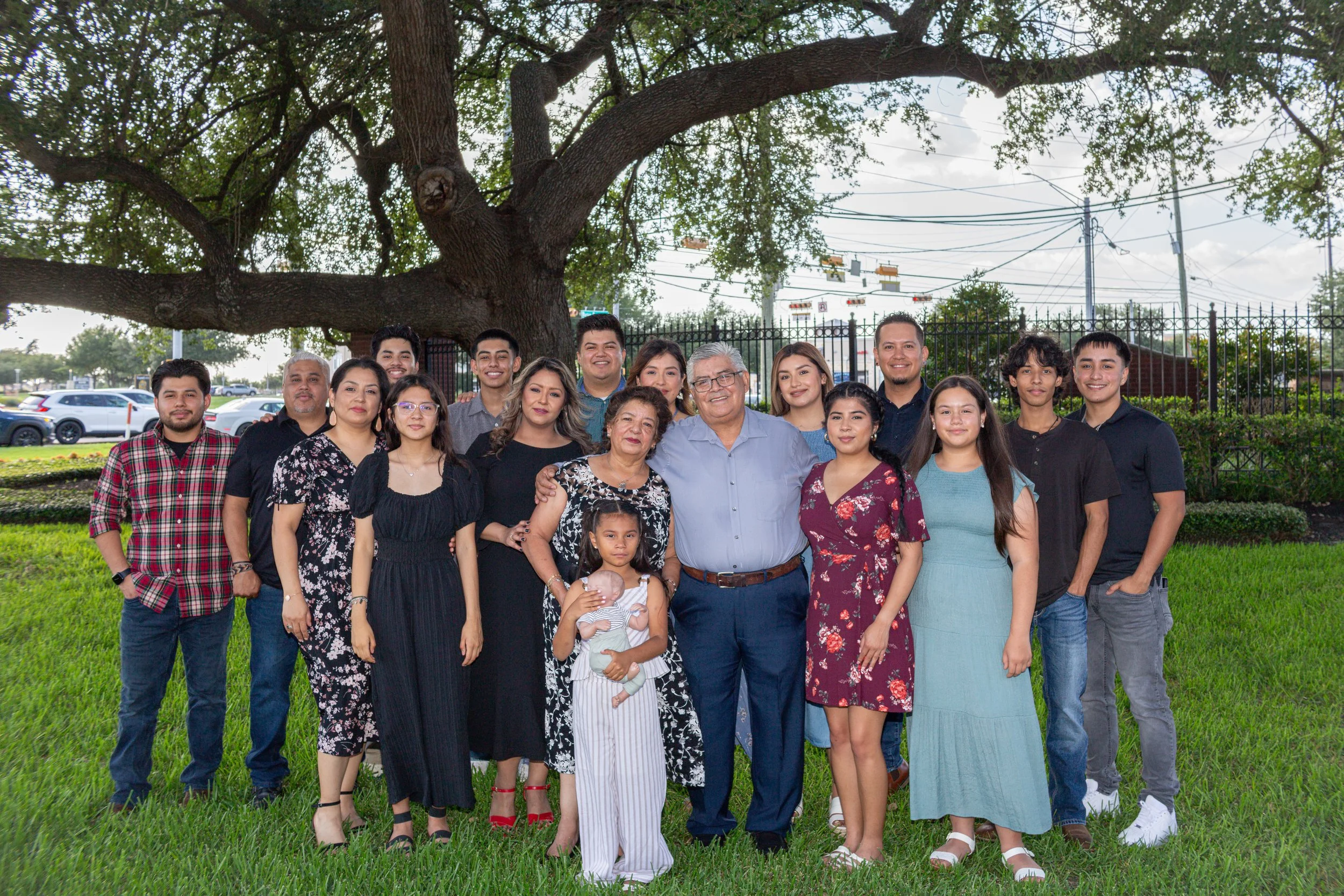 A large group of people, including adults and children, posing for a photo outdoors in front of a big tree with dense foliage. The group is standing on the grass, smiling, with a fence and parking lot visible in the background.
