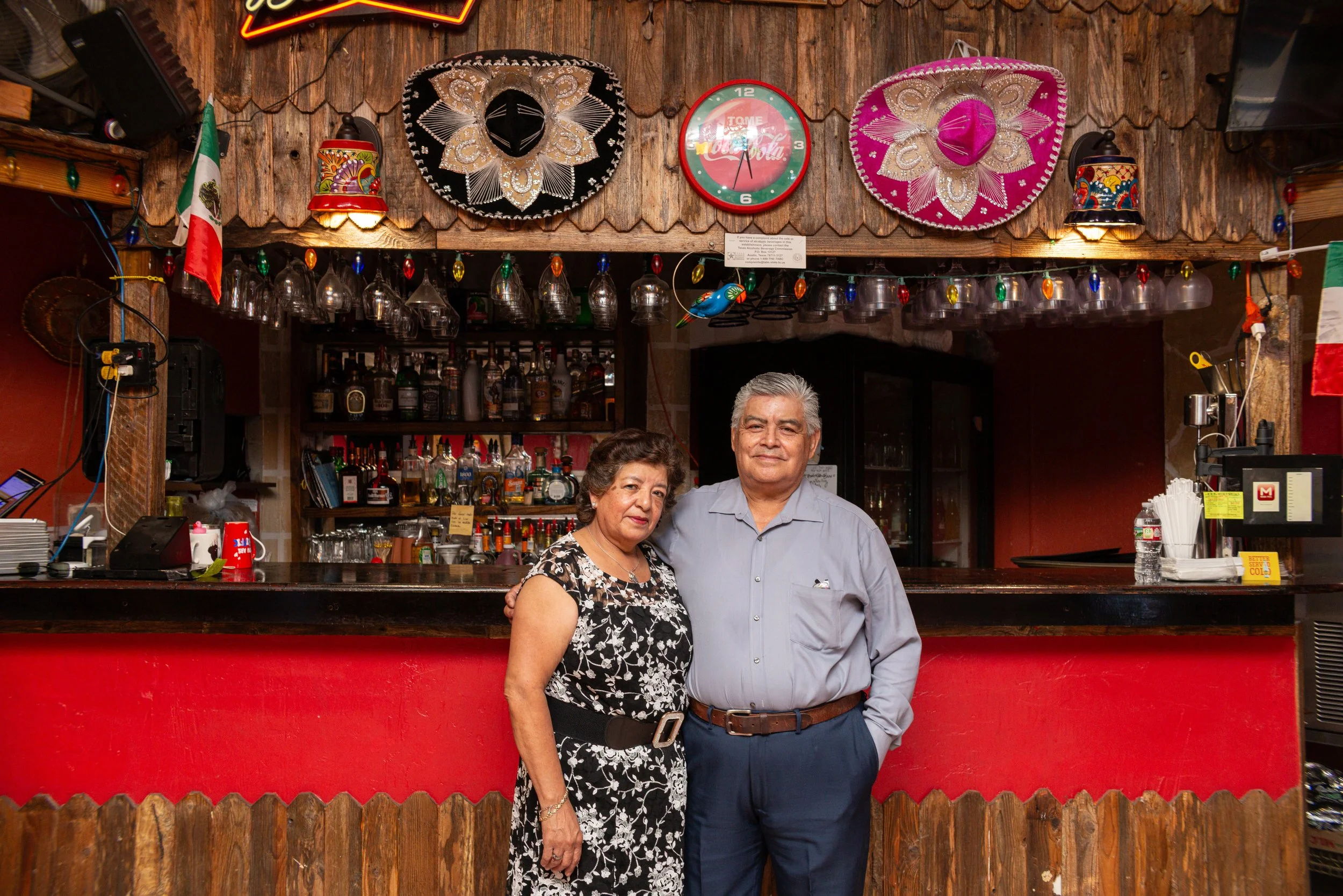 A middle-aged woman and man pose together behind a bar in a Mexican-themed restaurant or bar, decorated with sombreros, an American flag, a Coca-Cola clock, and colorful lights.