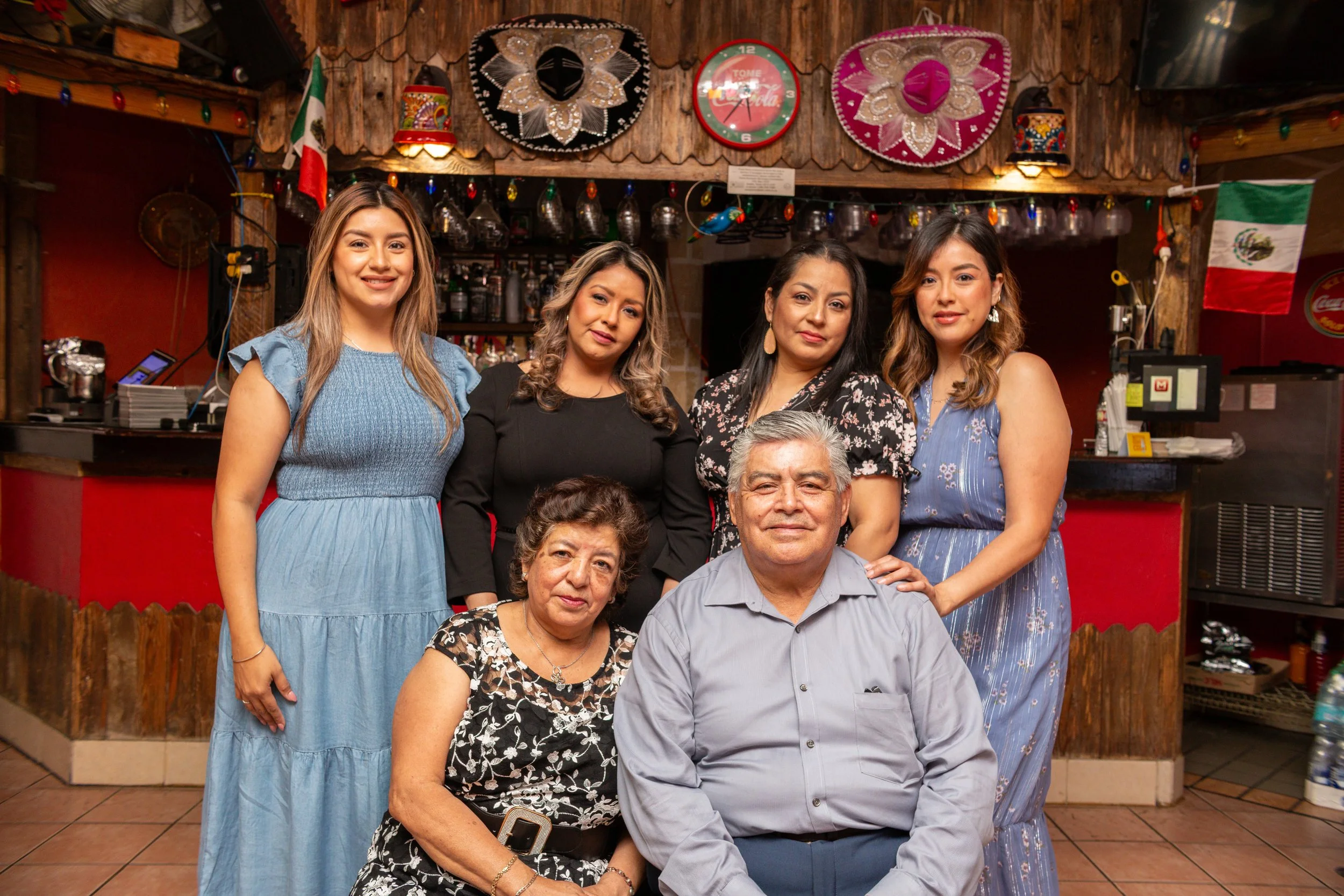 Family of six people in a restaurant with Mexican decorations, including sombreros and Mexican flags, smiling and posing for the camera.