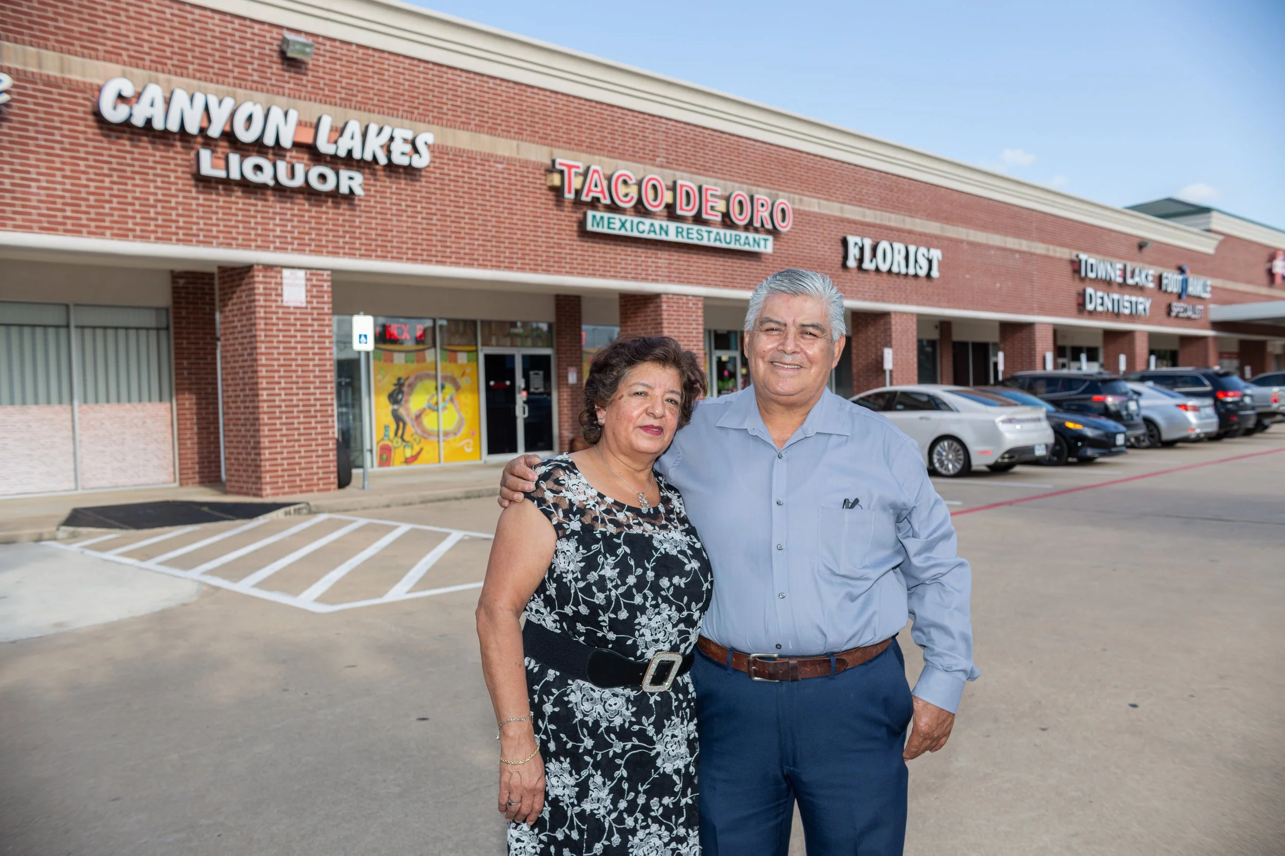 A smiling elderly man and woman standing together in front of a strip mall with various business signs including Taco Doro, Canyon Lakes Liquor, Florist, Towne Lake Food, and DENTISTRY, with parked cars in the background.