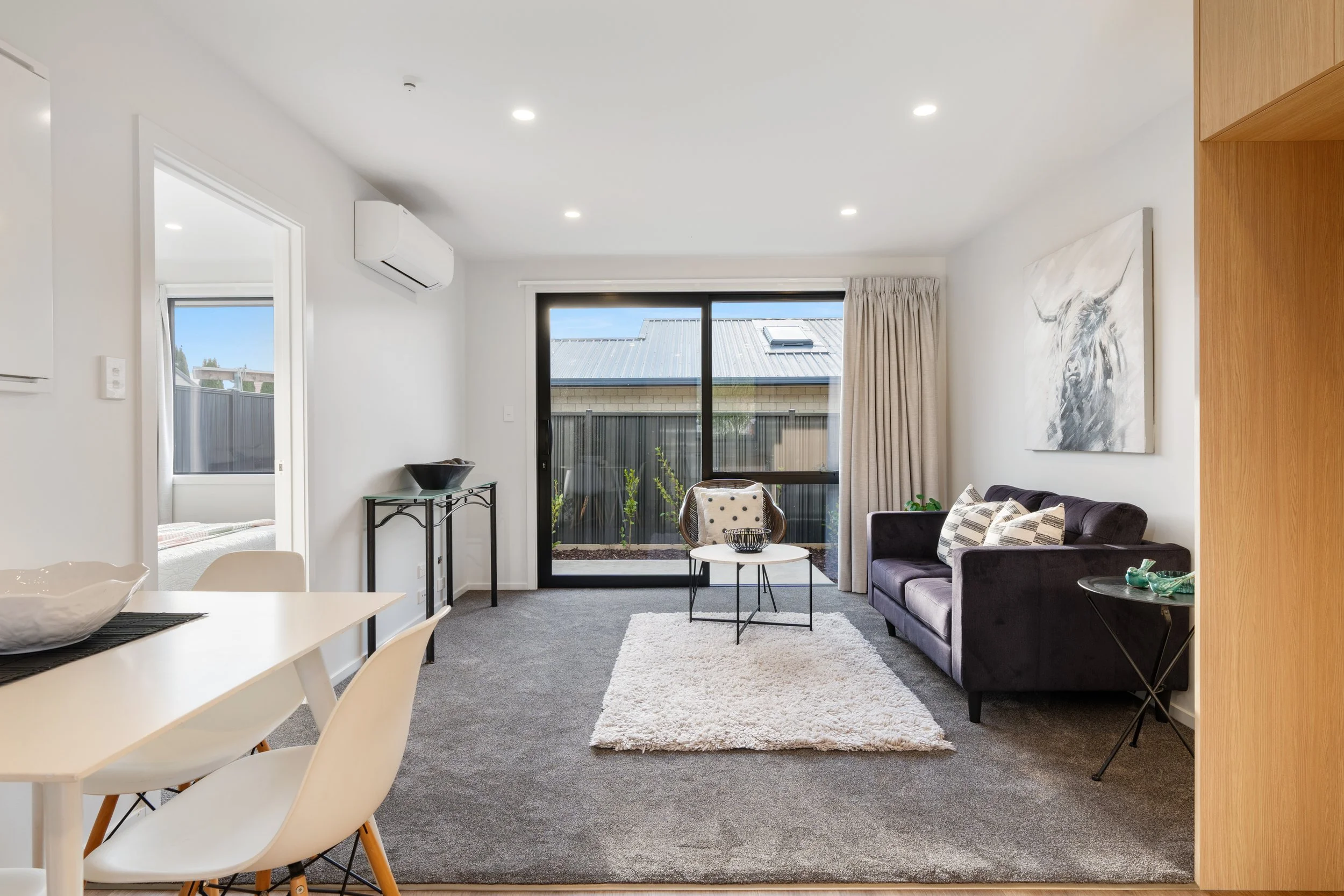 Bright living room with large sliding glass door, black sofa with patterned pillows, white shag rug, white walls, artwork, and beige curtains.