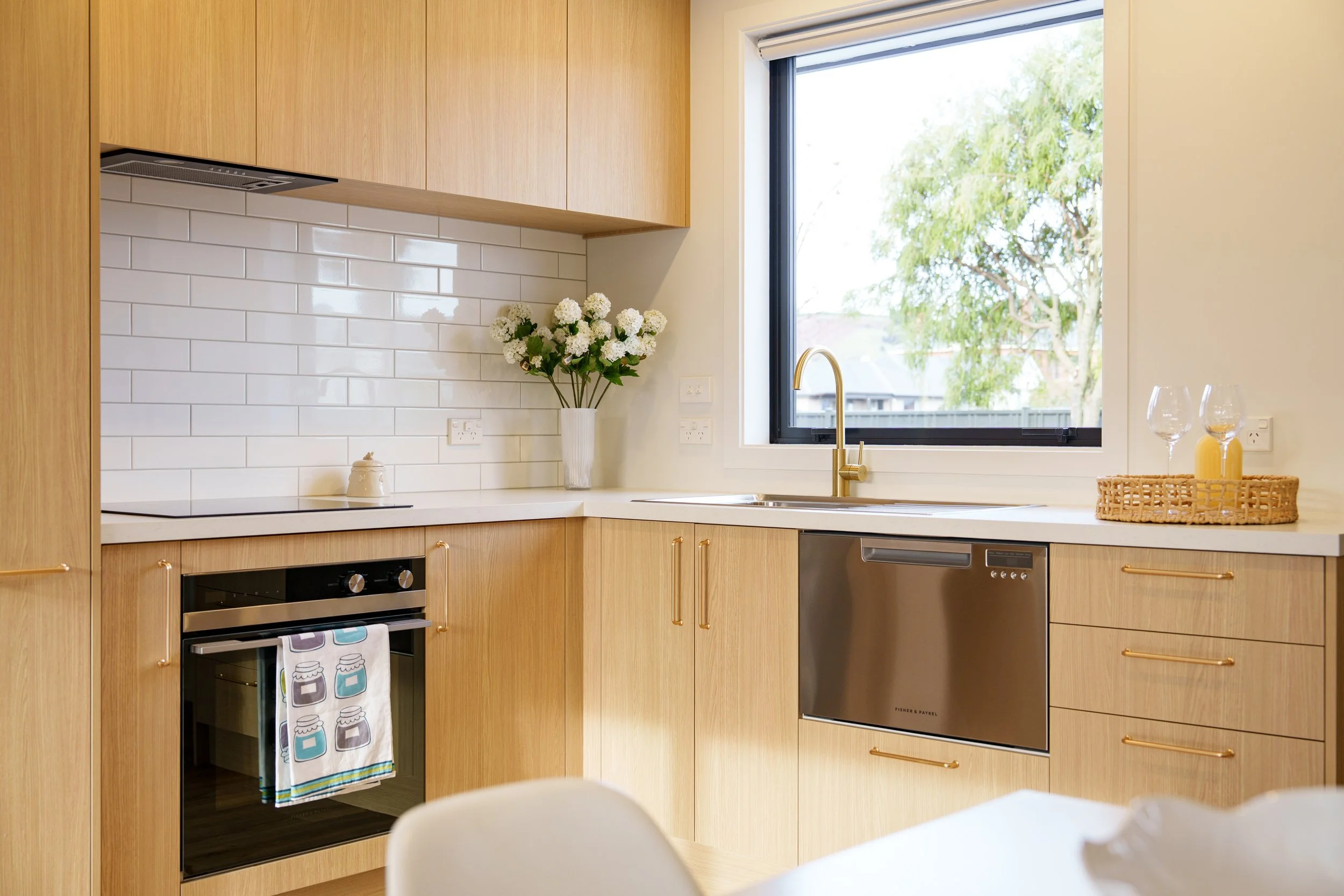 Modern kitchen with wooden cabinets, white subway tile backsplash, and a large window with a view of trees. Contains stainless steel oven, dishwasher, gold faucet, and a vase of white flowers.