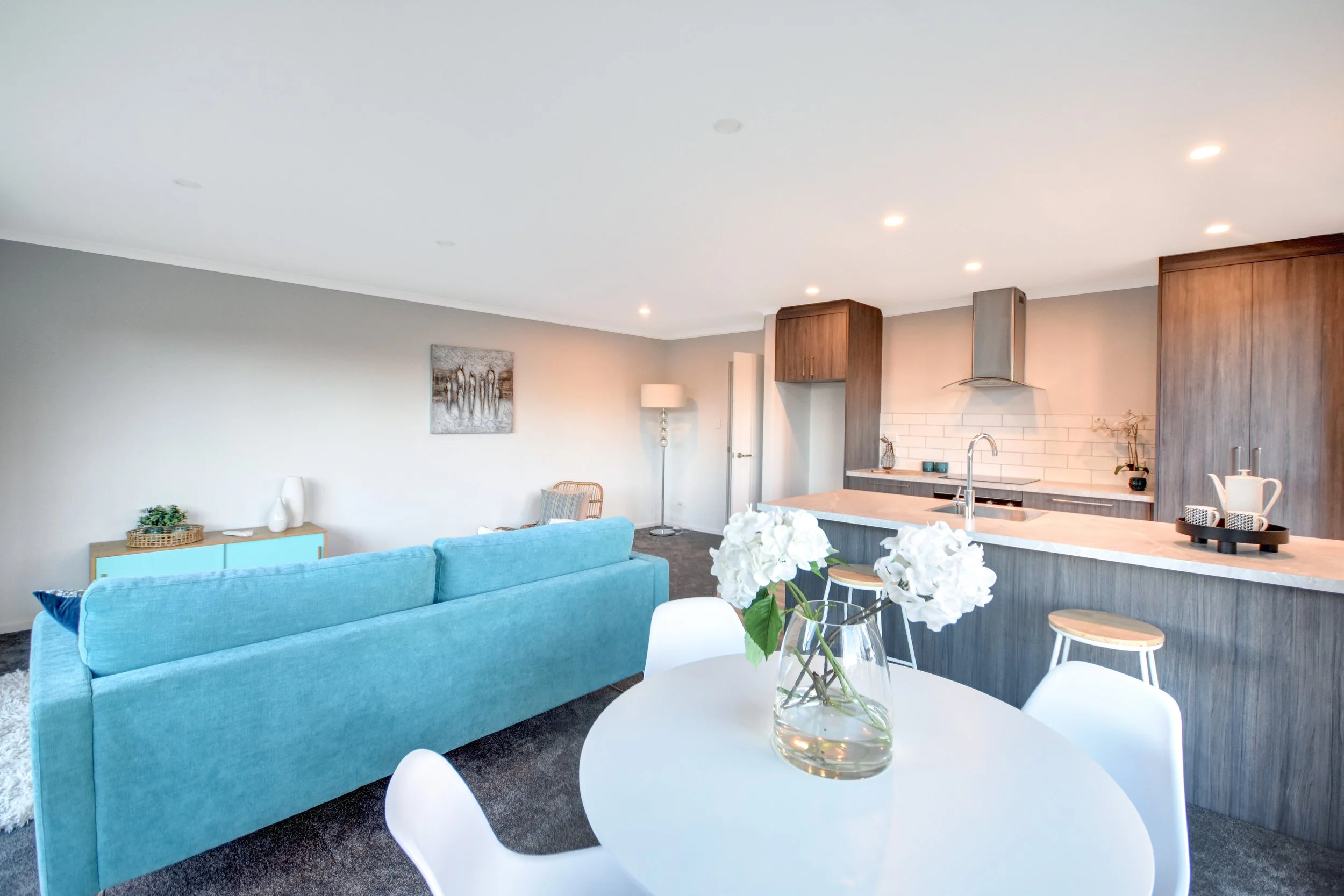 Open-concept living room and kitchen with blue sofa, white dining table with flowers, wooden cabinets, and modern kitchen appliances.