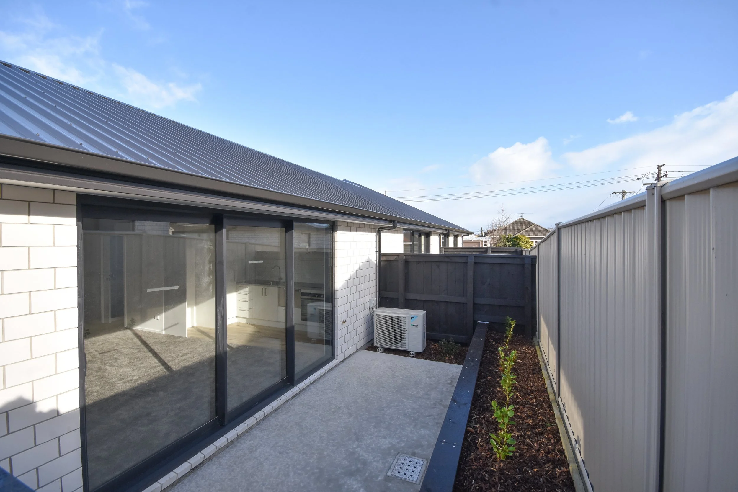 Small backyard patio with concrete flooring, a garden bed with small green plants, enclosed by a beige metal fence, featuring a modern house with large glass sliding doors, white brick exterior, and a solar panel roof.
