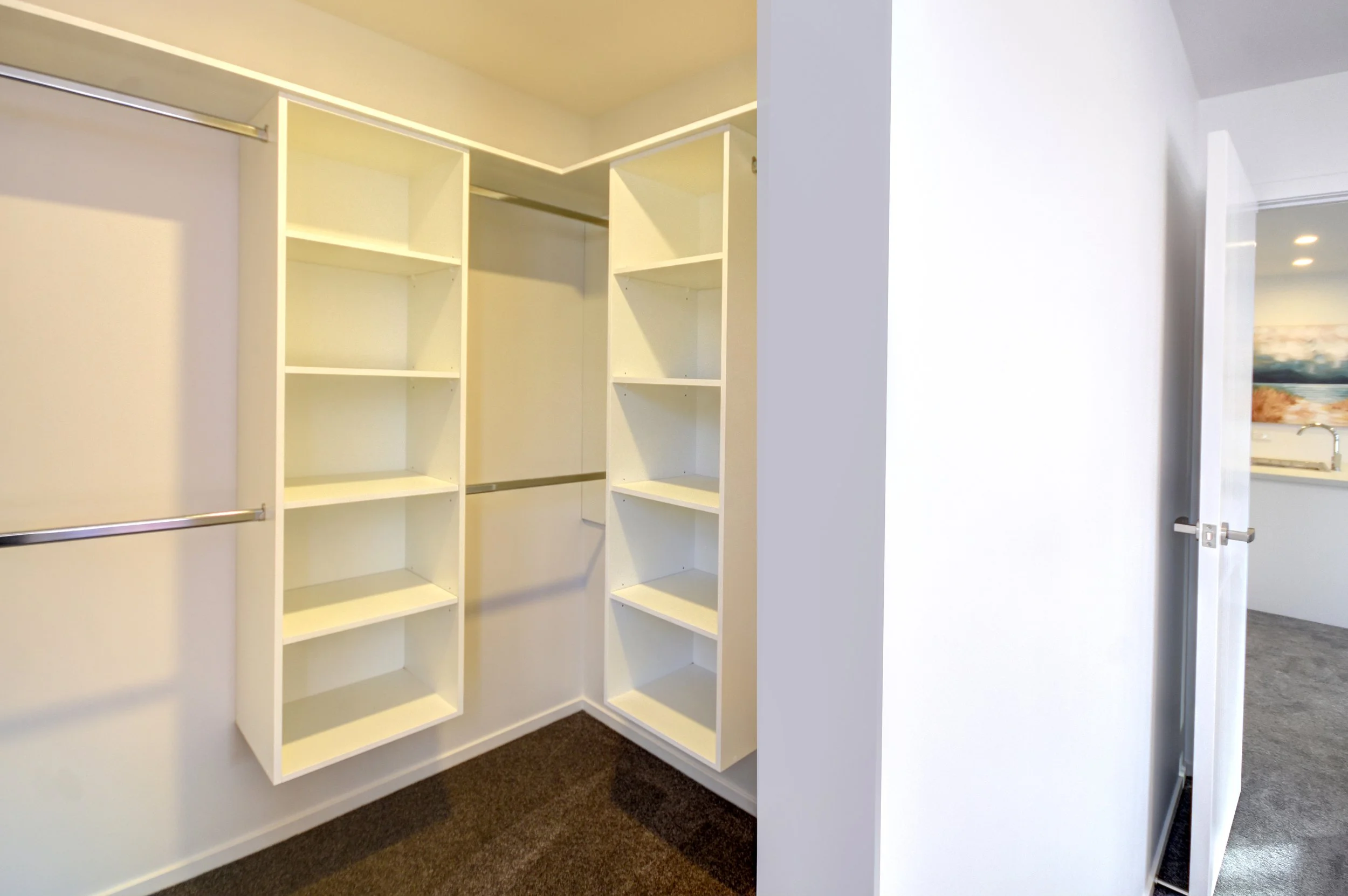 Empty walk-in closet with white shelving units and hanging rods, adjacent to a room visible through an open door.