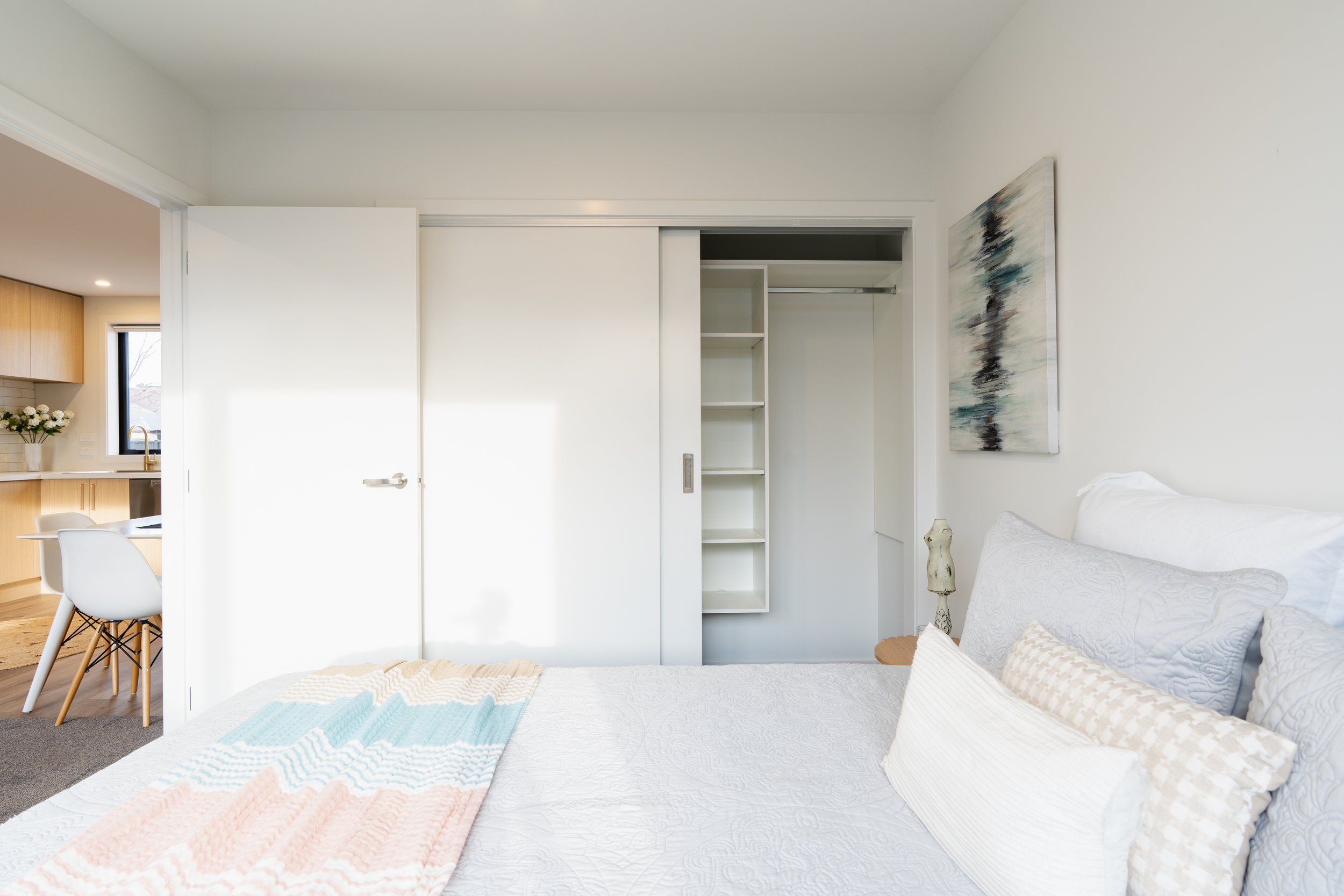 A bedroom with a bed, pillows, a painting on the wall, and an open closet with shelves. Adjacent to the bedroom is a dining area with a white chair, a table, and a kitchen visible in the background.