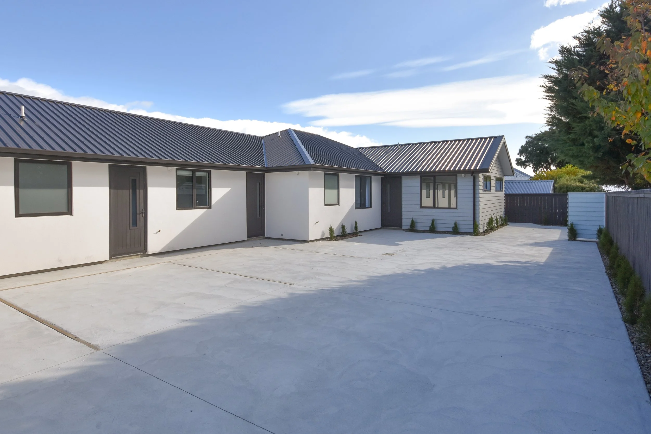 Modern house with metal roofing and a large concrete patio, surrounded by a wooden fence and some small shrubs.