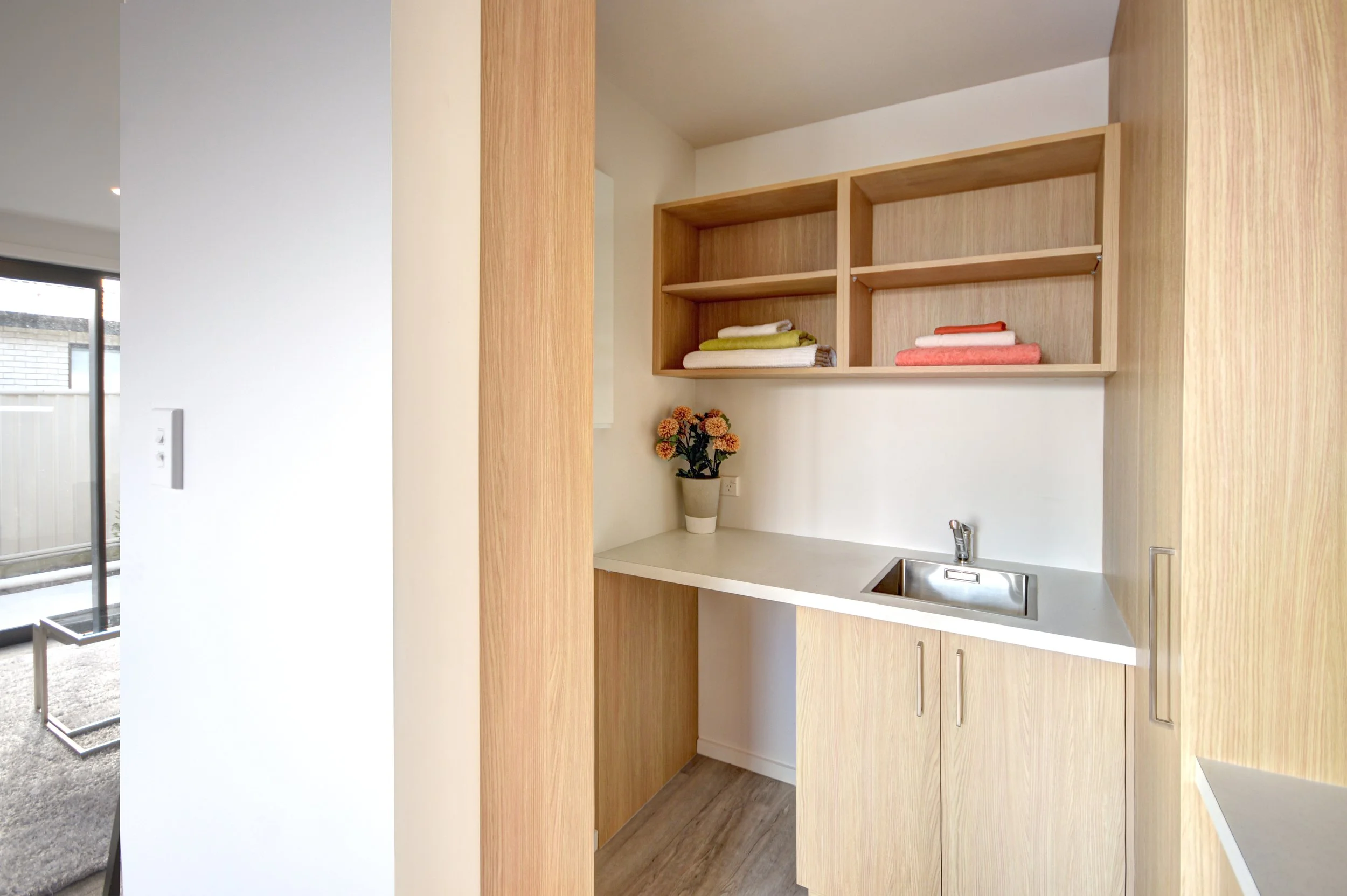Small kitchenette area with light wood cabinets, open shelving with neatly folded towels, a white countertop, a small sink with a chrome faucet, and a potted plant with orange flowers.