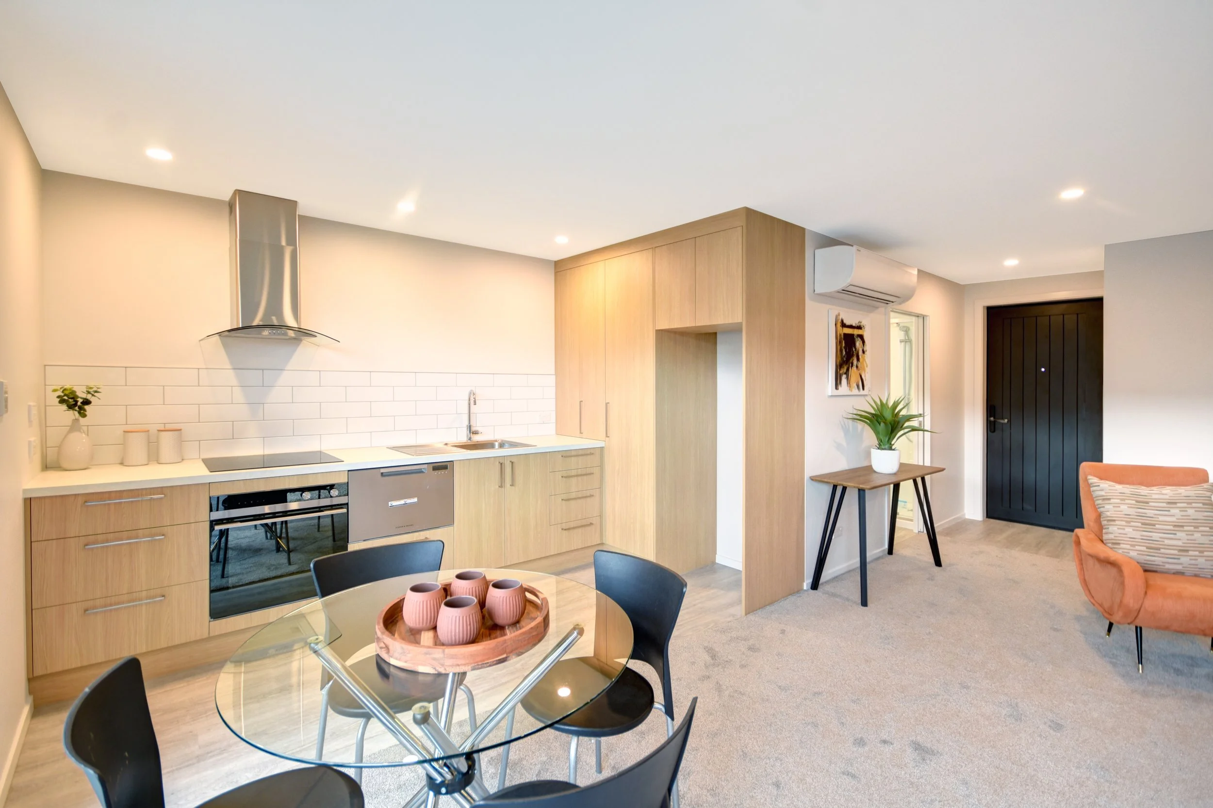 Open kitchen and living room with wood cabinets, white subway tile backsplash, black appliances, round glass dining table with five black chairs, orange sofa, and green plants.