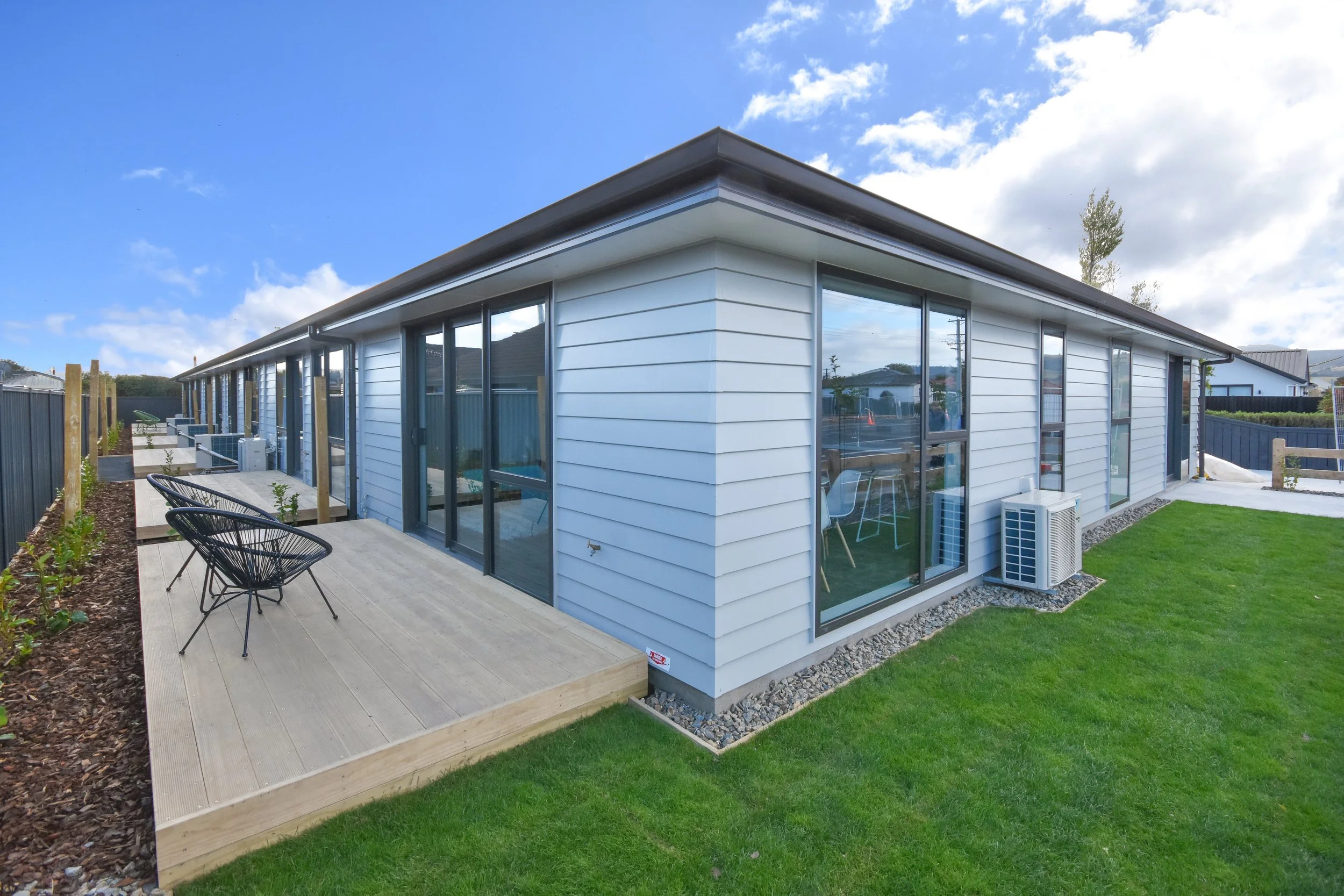 Exterior view of a modern, single-story residential building with large glass sliding doors and a wooden deck with black outdoor chairs, surrounded by a green lawn and a dark gray fence under a partly cloudy sky.