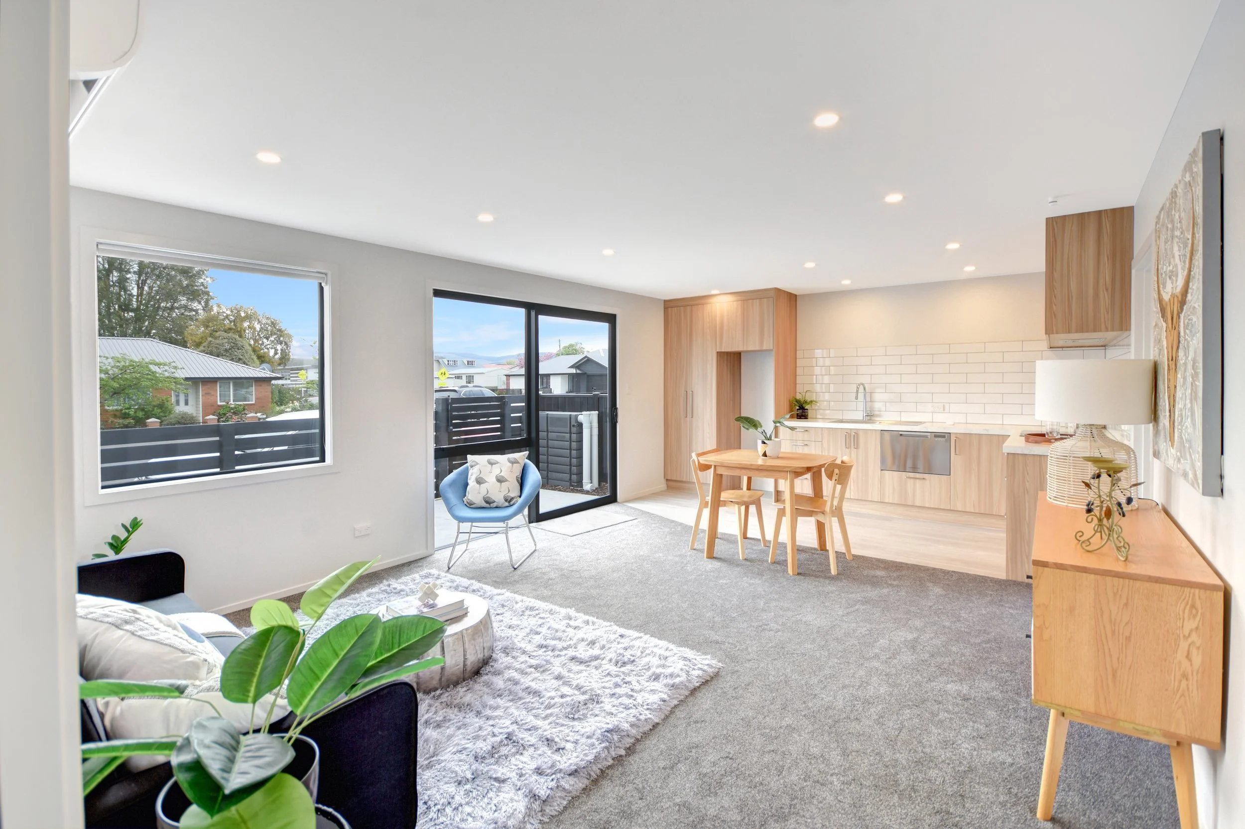 Open-concept living room with large windows and sliding door, contemporary furniture, a cozy area rug, a sectional sofa, a small round coffee table, potted plants, and a kitchen with wooden cabinets and a dining table.