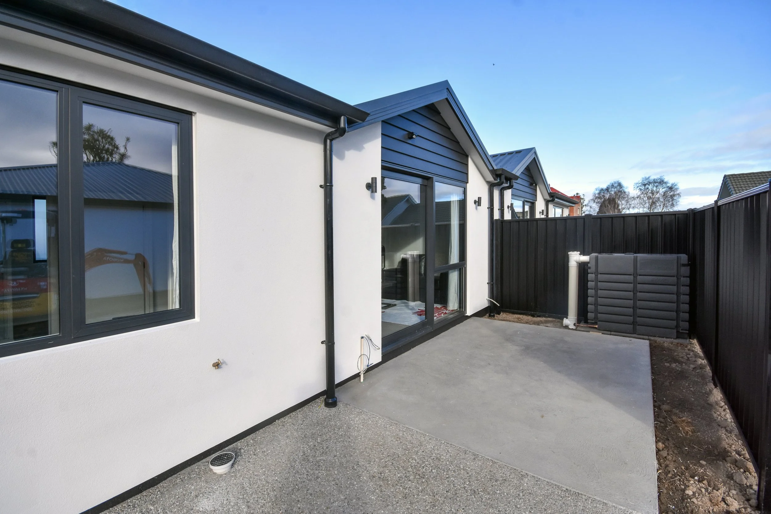 Newly built house exterior with white walls, black window frames, and a small concrete patio area enclosed by a black wooden fence.
