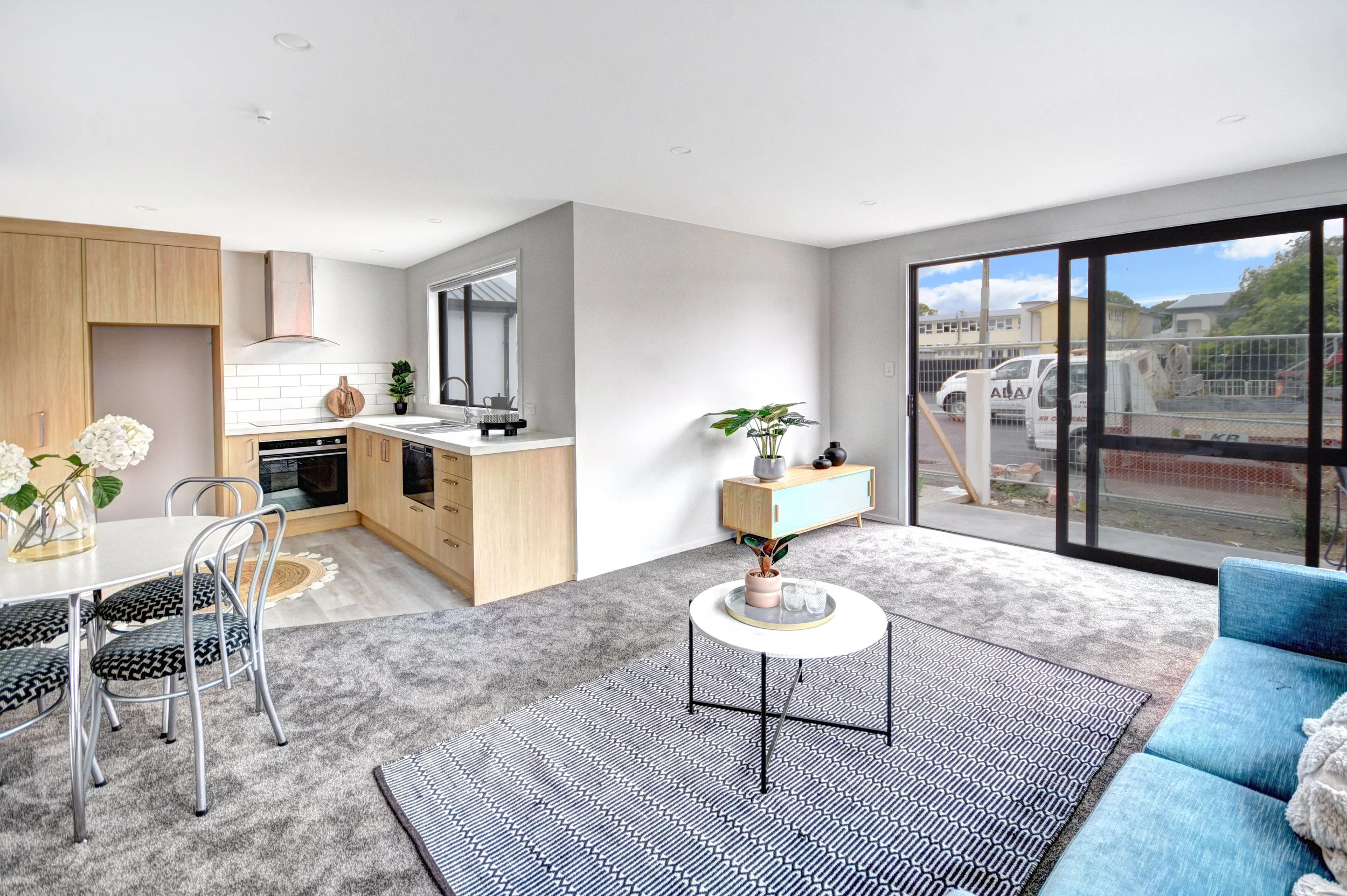 Open-concept living room and kitchen in a modern home with large sliding glass doors, blue sofa, white coffee table, dining table with chairs, and minimalistic decor.