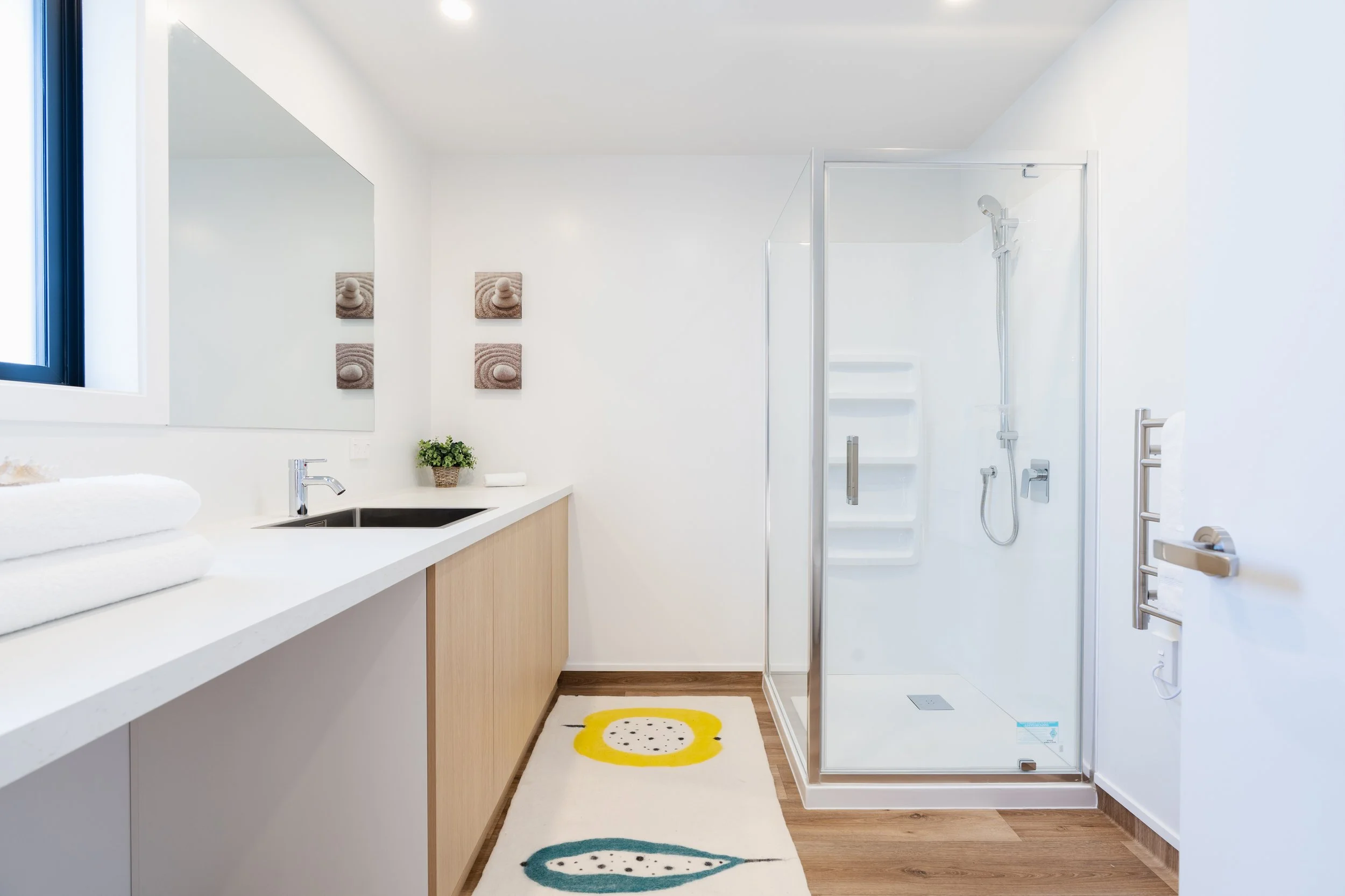 Modern bathroom with white walls, a walk-in shower with glass door, wooden floor, and vanity with a white countertop, black sink, and stacked towels. There is a mirror above the vanity, wall art, a small plant, a yellow and teal patterned rug, and a 
