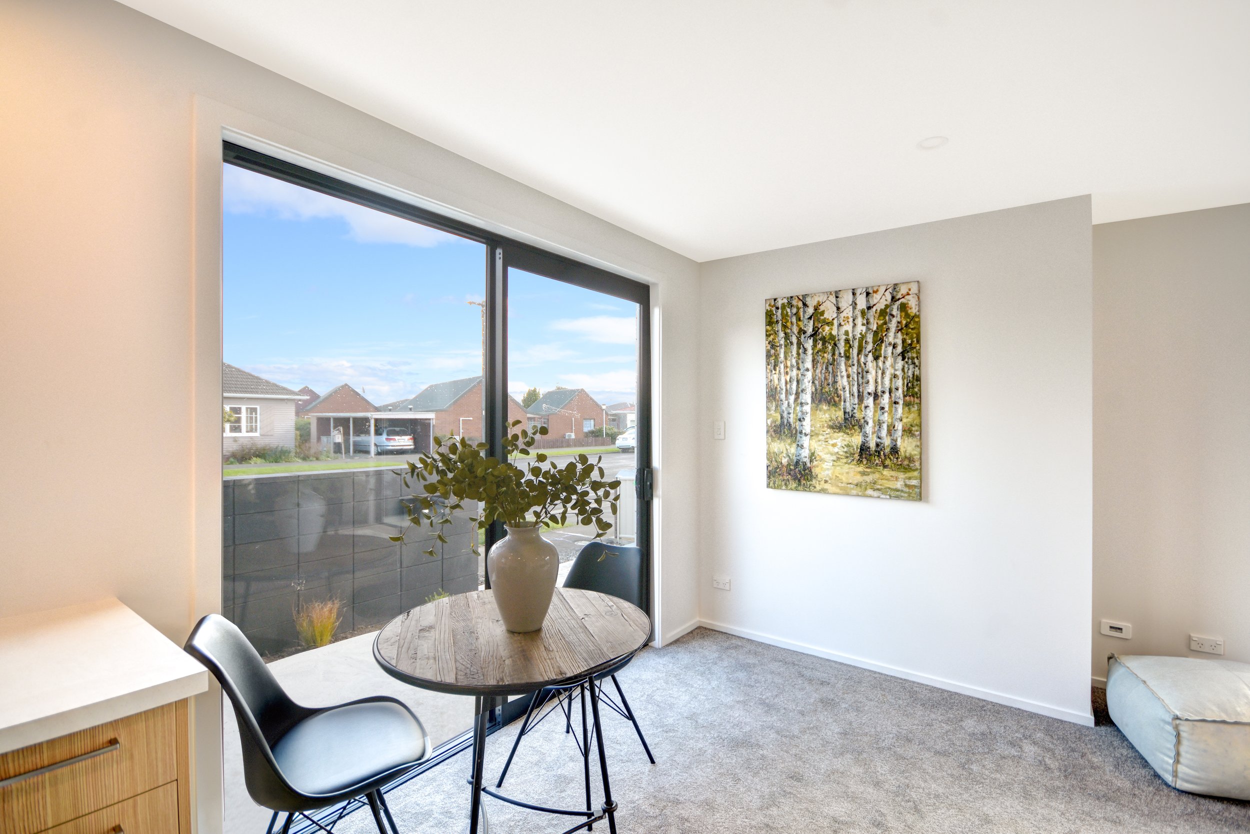 Interior view of a room with a small round wooden table, a vase with green foliage, a black chair, a large window showing houses outside, a framed painting of a birch forest on the wall, and a light-colored floor