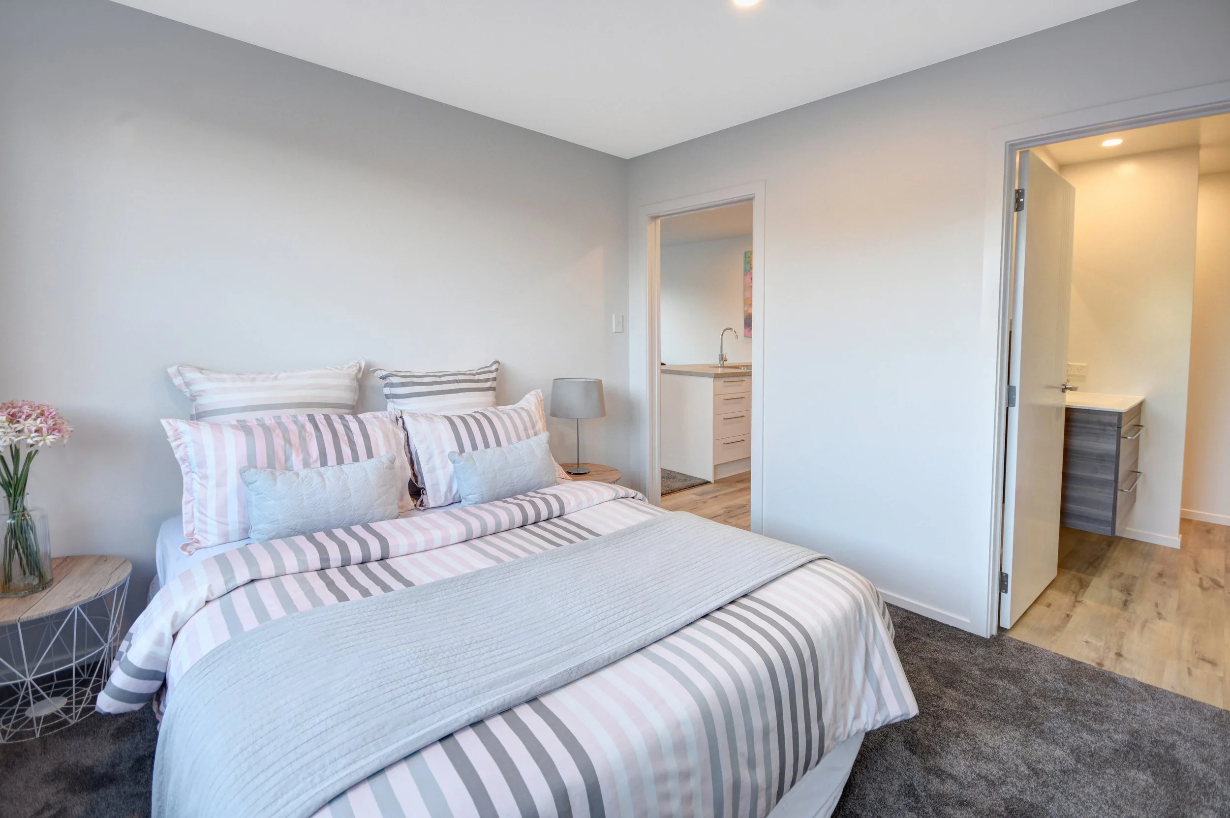 A bedroom with white walls and a bed with striped bedding and multiple pillows. A bedside table with a glass vase of pink flowers and a small gray lamp. An open door reveals a hallway and laundry area with wooden floors and white cabinets.