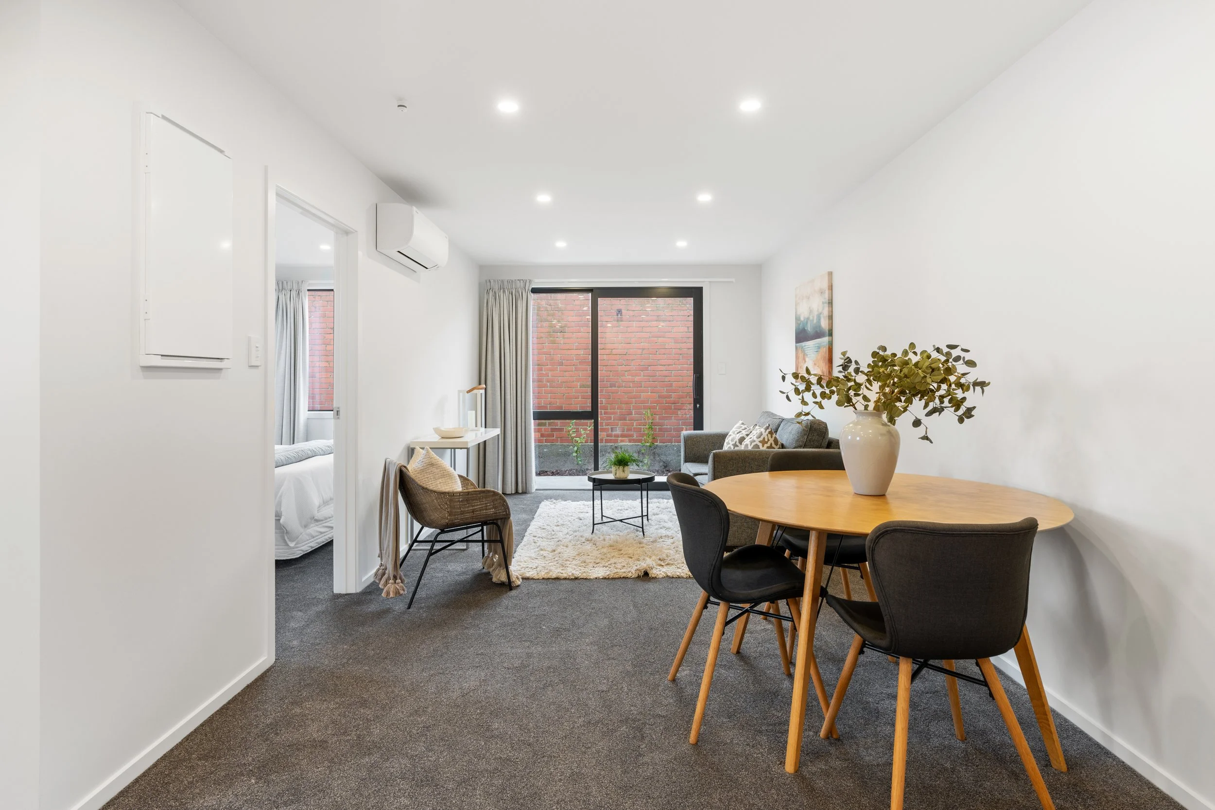 Living room with gray sofa, round wooden dining table with black chairs, sliding glass door with brick wall outside, white walls, beige curtains, and artwork on wall.