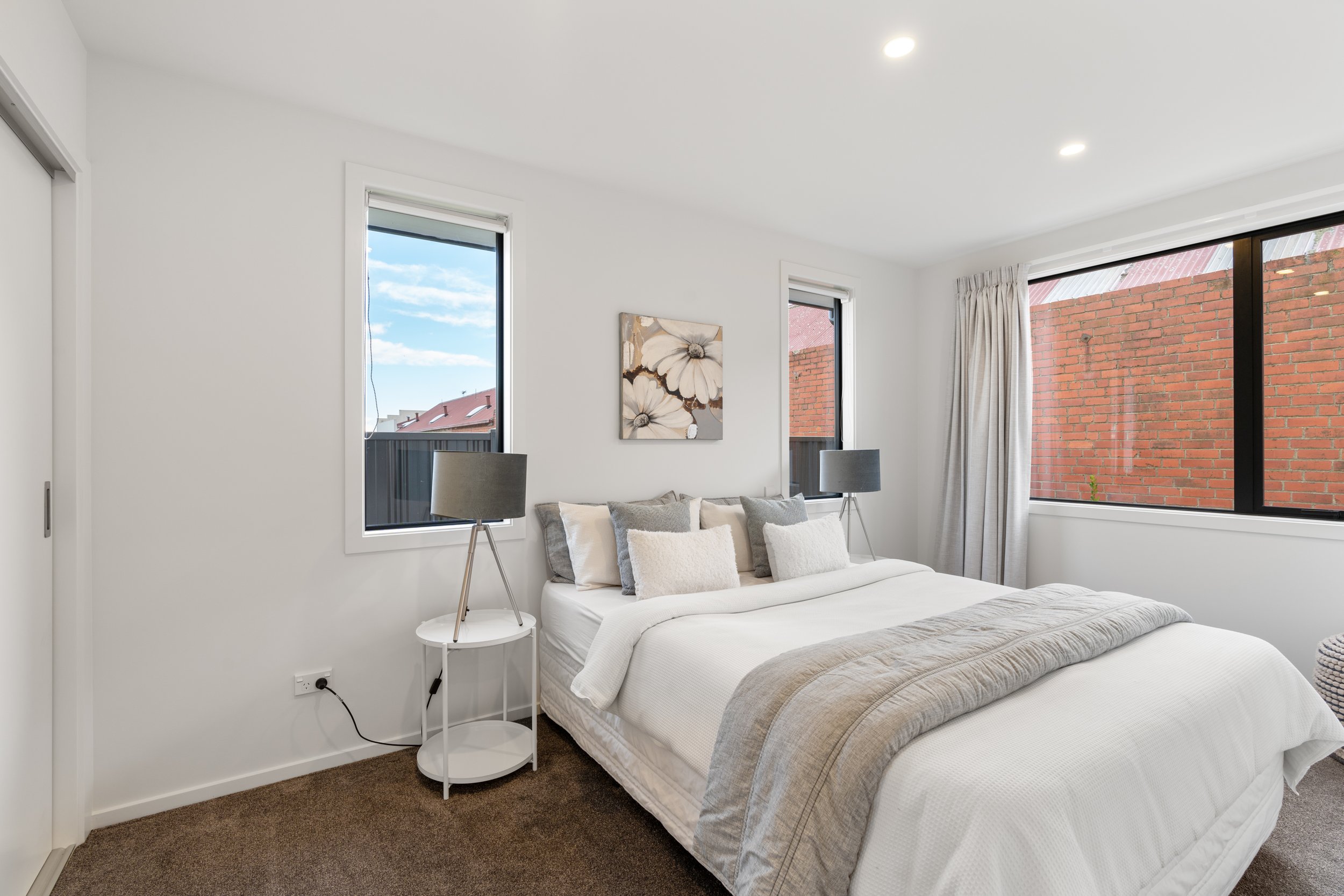 A modern bedroom with white walls, a white bed with multiple gray and white pillows, two side tables with table lamps, and large windows with a view of brick walls outside. A framed floral artwork hangs above the bed.