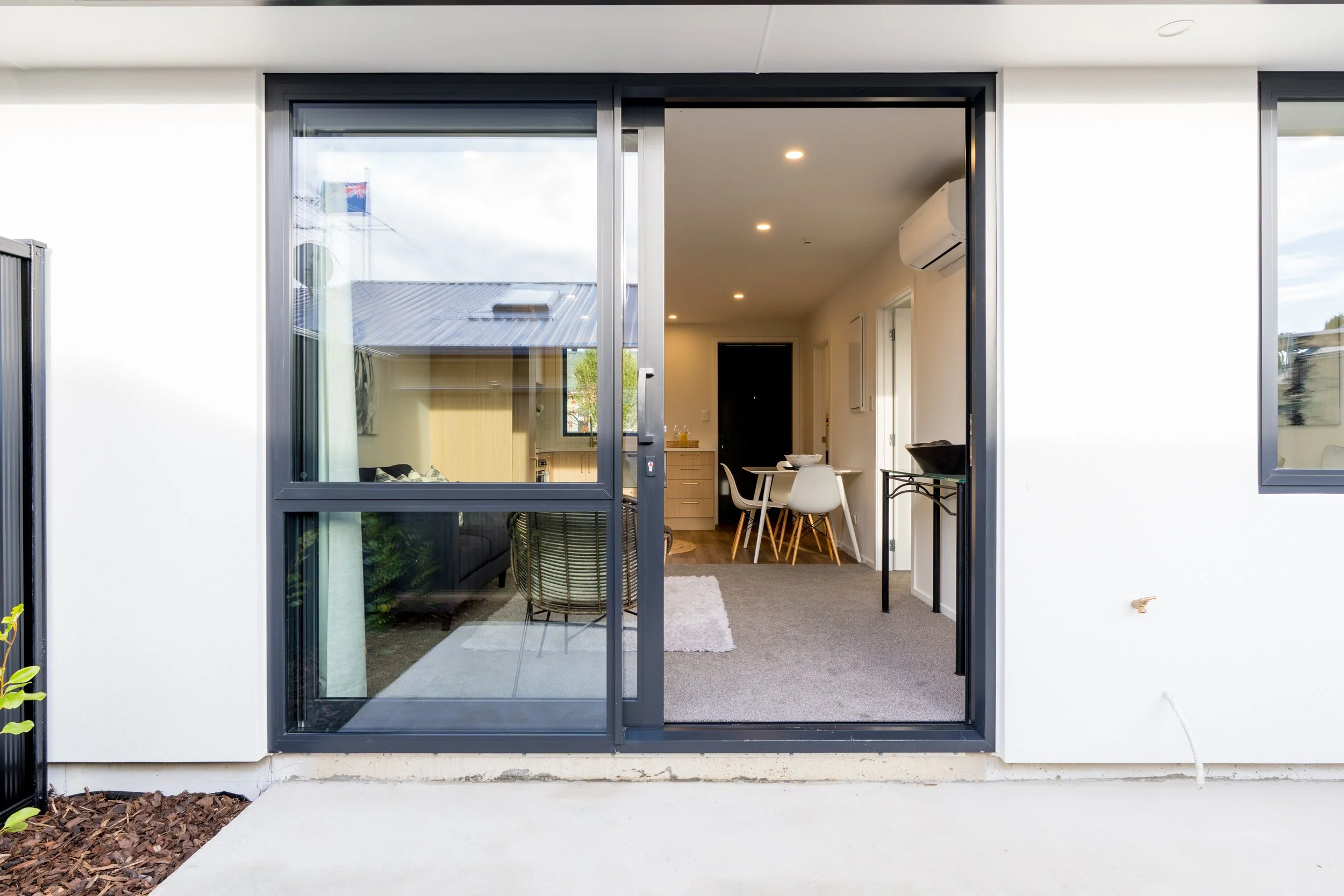 Open sliding glass door leading into a modern living room and dining area with white walls, carpeted floor, dining table with chairs, and an air conditioning unit on the wall.