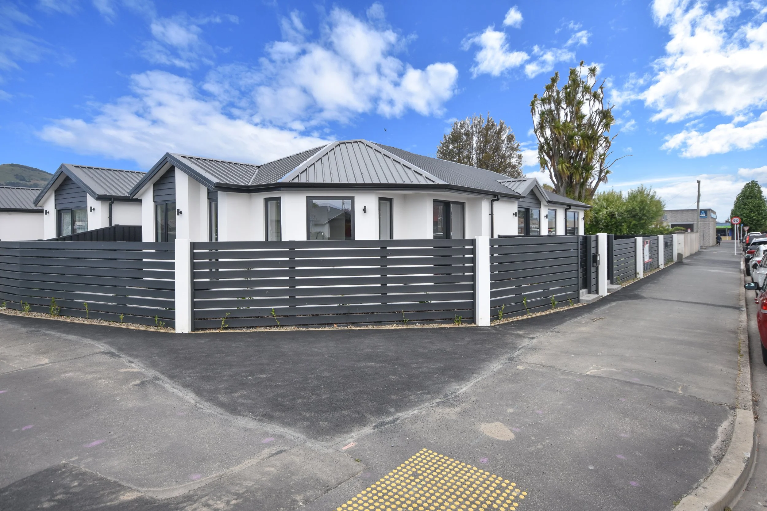 Modern white house with black roof and fencing, accessible sidewalk, cars parked on the street, trees, cloudy blue sky.