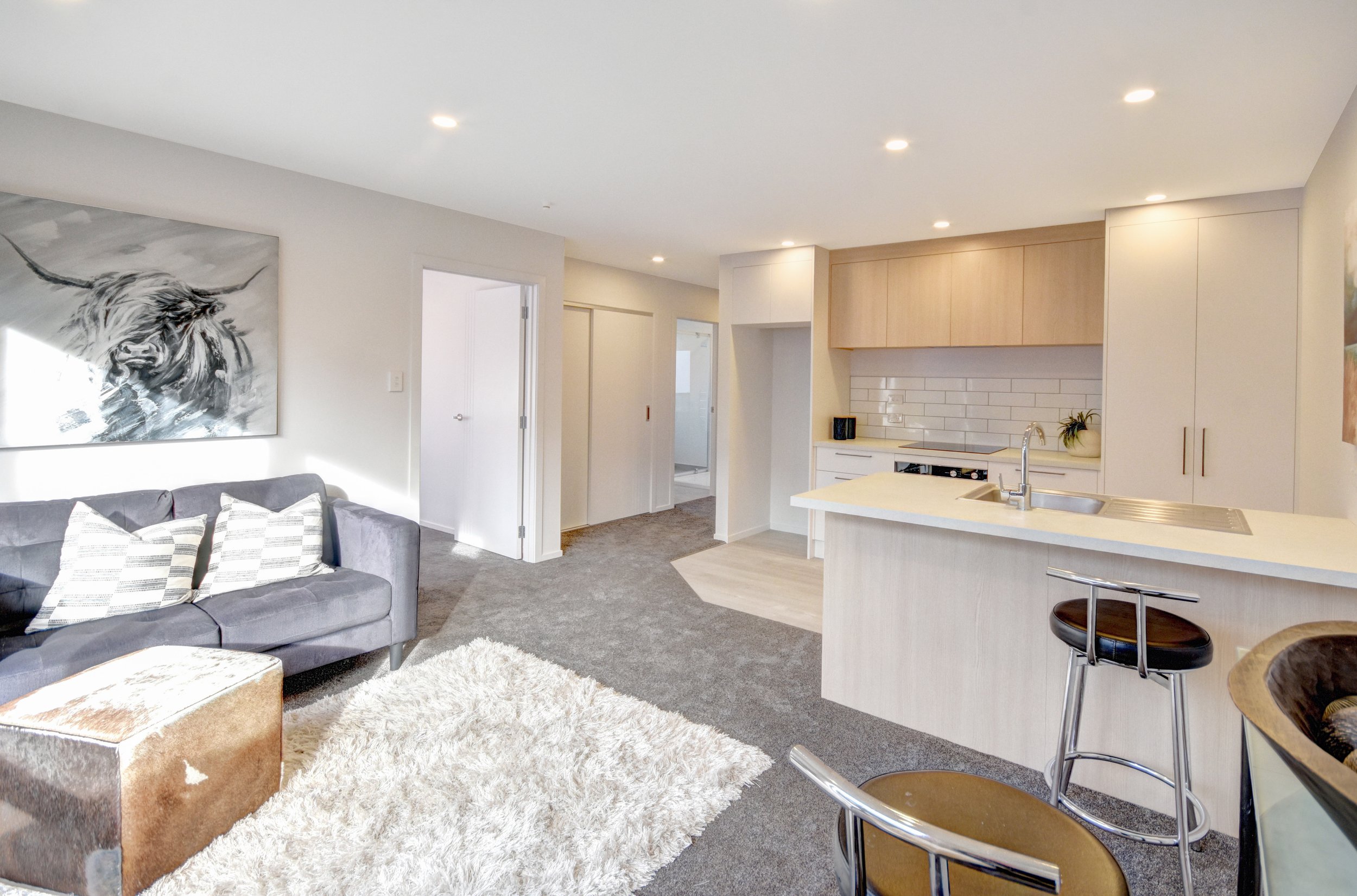 Open-concept living room and kitchen with gray sofa, white shag rug, abstract black and white animal painting, white cabinetry, and a kitchen island with bar stools.