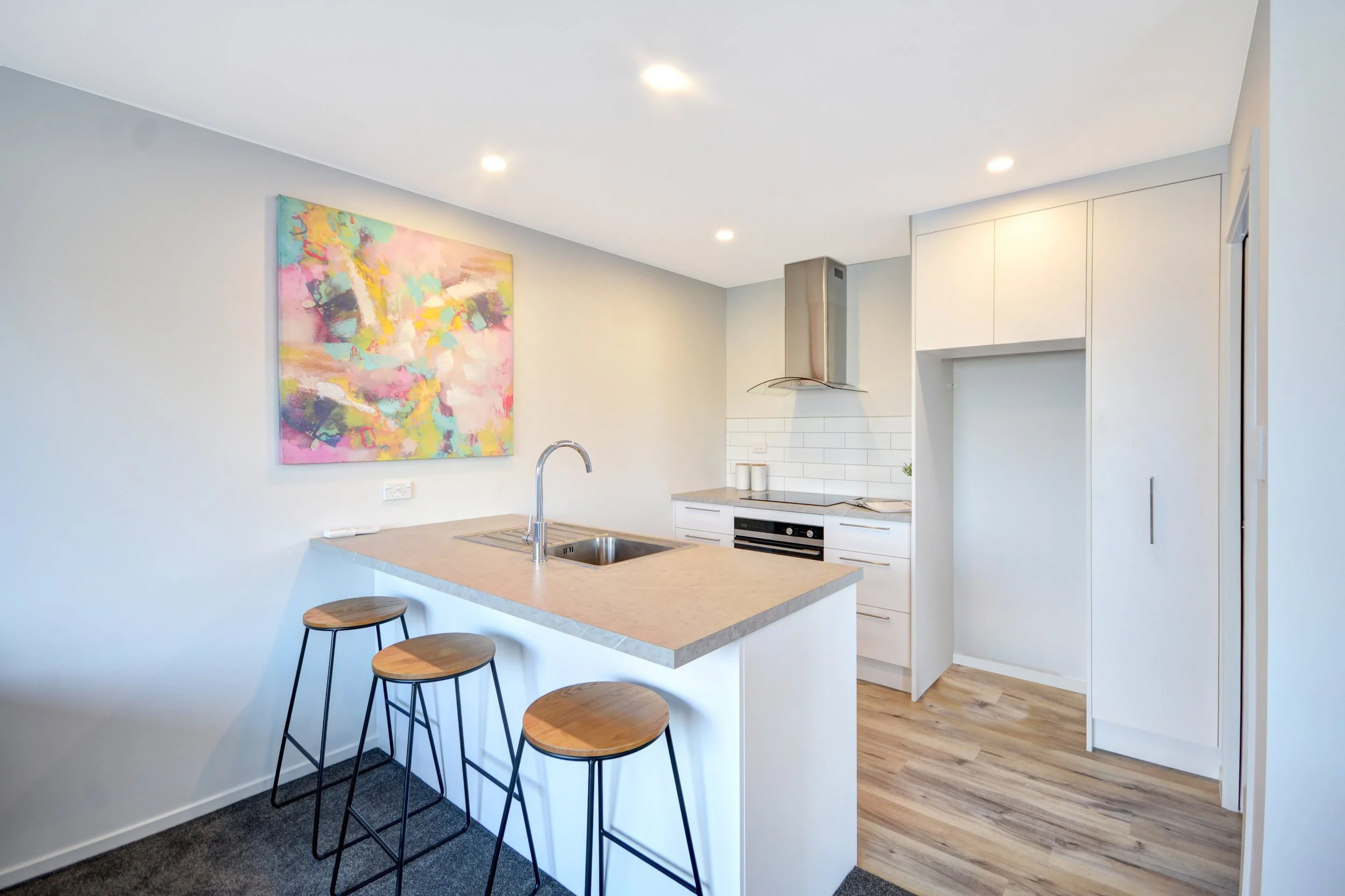 Modern kitchen with white cabinets, a gray countertop island with a sink and three barstools, an abstract colorful painting on the wall, and a wood floor.
