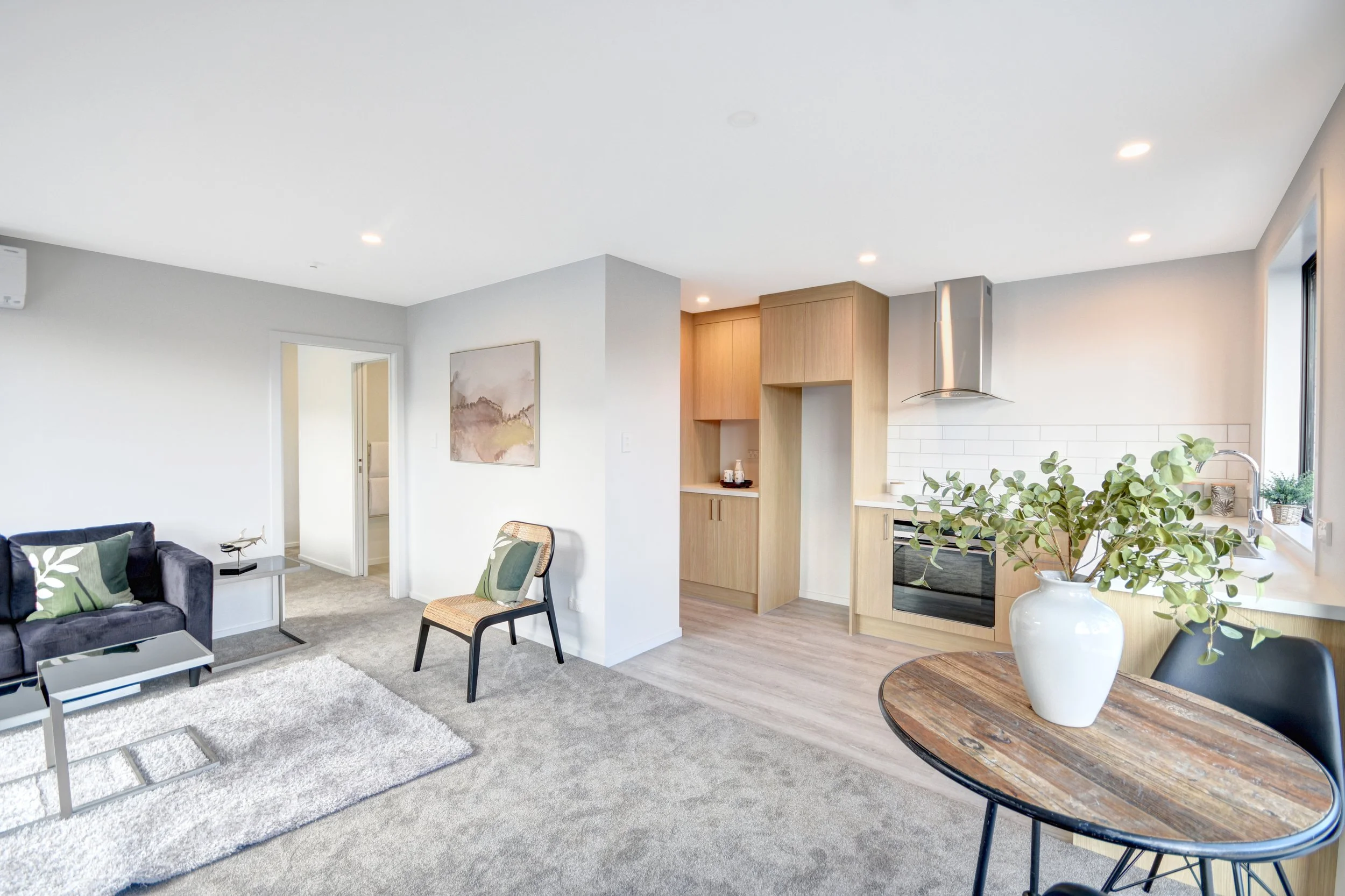 Open-concept living room and kitchen with light-colored walls, a dark sofa with green pillows, a wooden dining table with a white vase and greenery, and modern kitchen appliances and cabinetry.