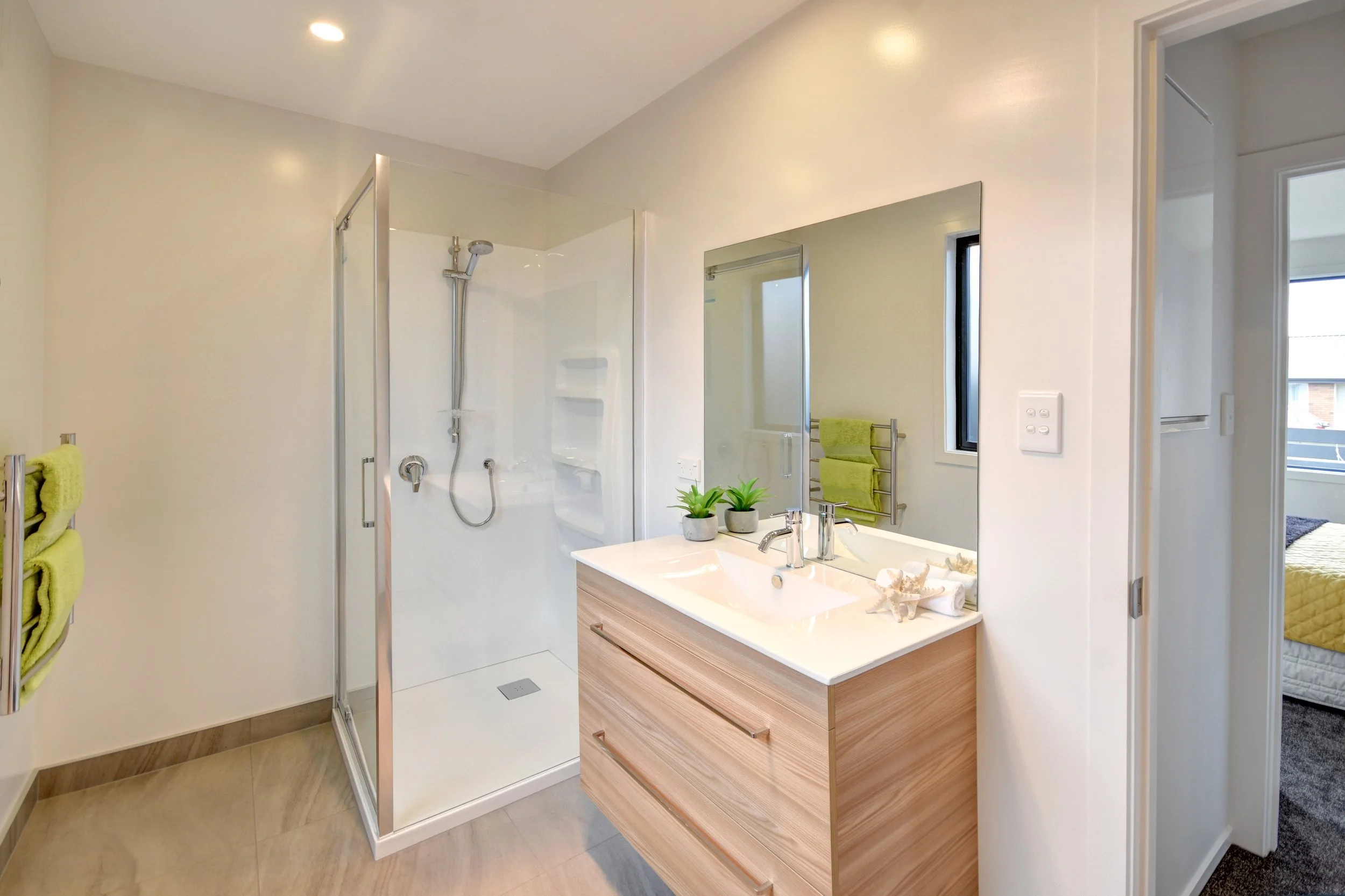 Modern bathroom with a walk-in shower, a wooden vanity with a white countertop, a large mirror, and green towels hanging on a heated towel rack. A bedroom with a window and a yellow bedspread is visible through an open door.