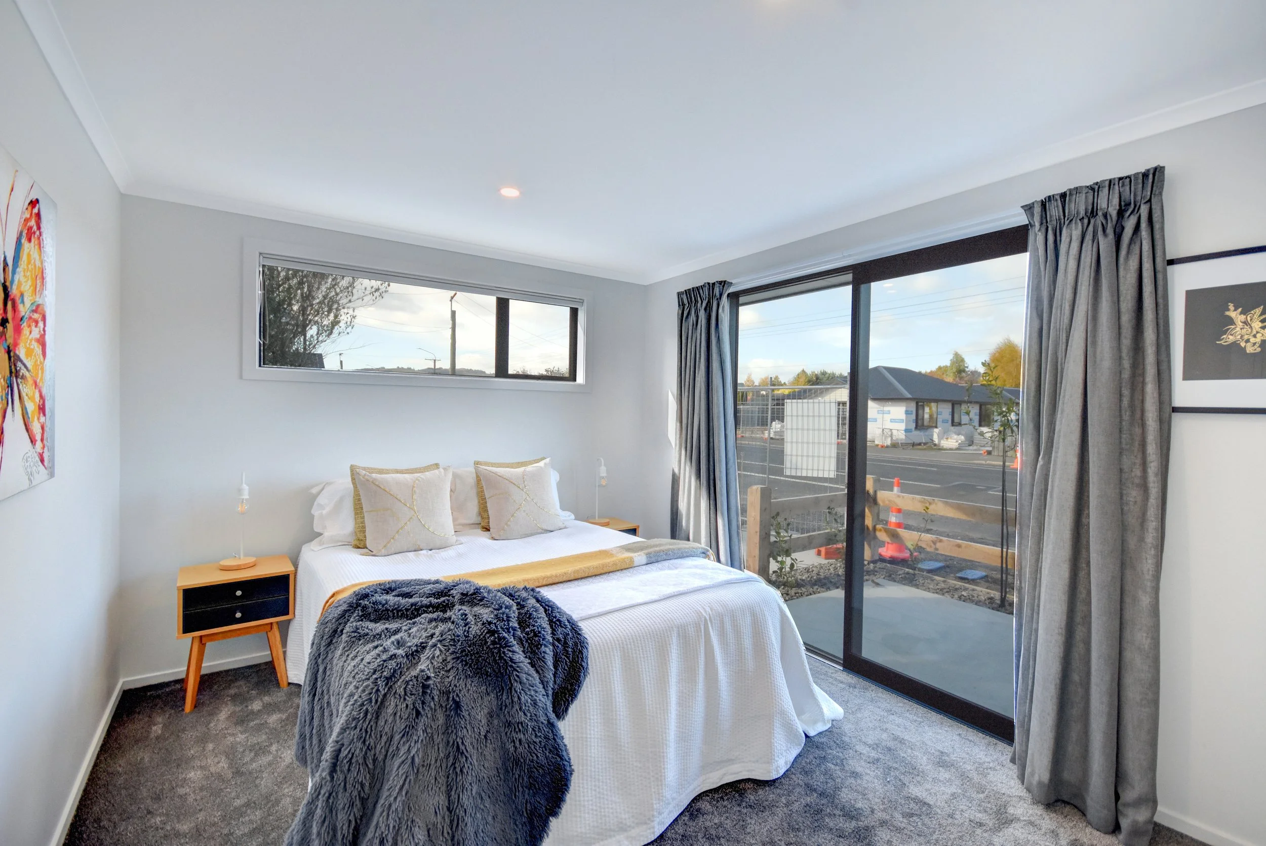 Bedroom with white bedding, pillows, dark gray blanket, nightstand, artwork, large window with curtains, sliding glass door leading outside, views of a street under construction