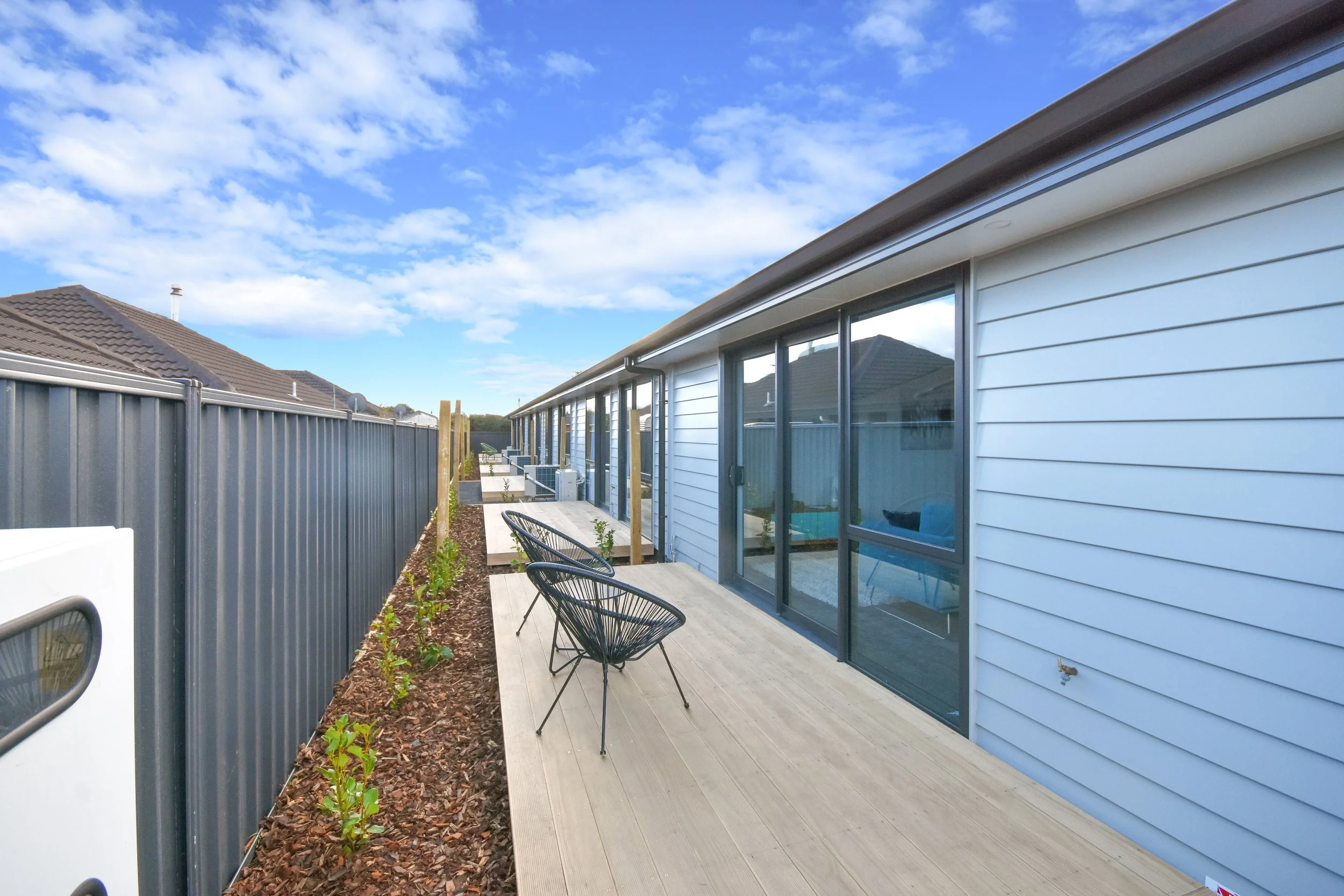 Backyard patio area with a wooden deck, black wire chairs, sliding glass doors, a gray metal fence, and newly planted bushes, under a partly cloudy sky.