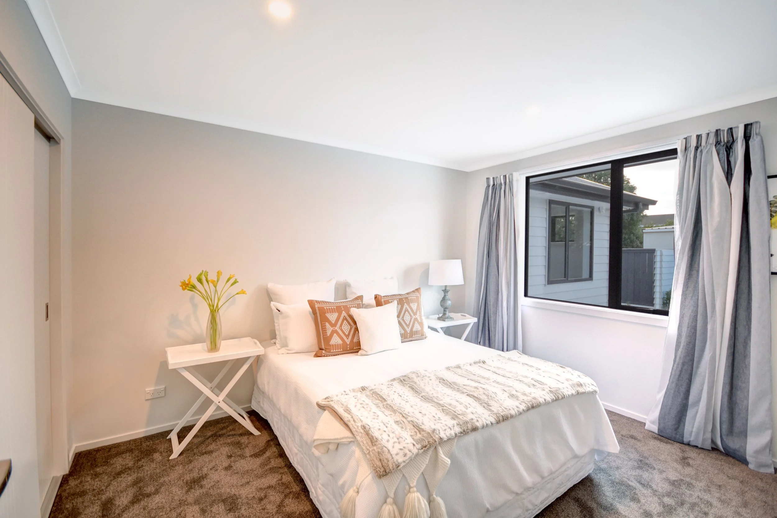 A bedroom with a bed, white bedding, decorative pillows, a bedside table with a lamp, a vase of flowers, and a window with curtains.