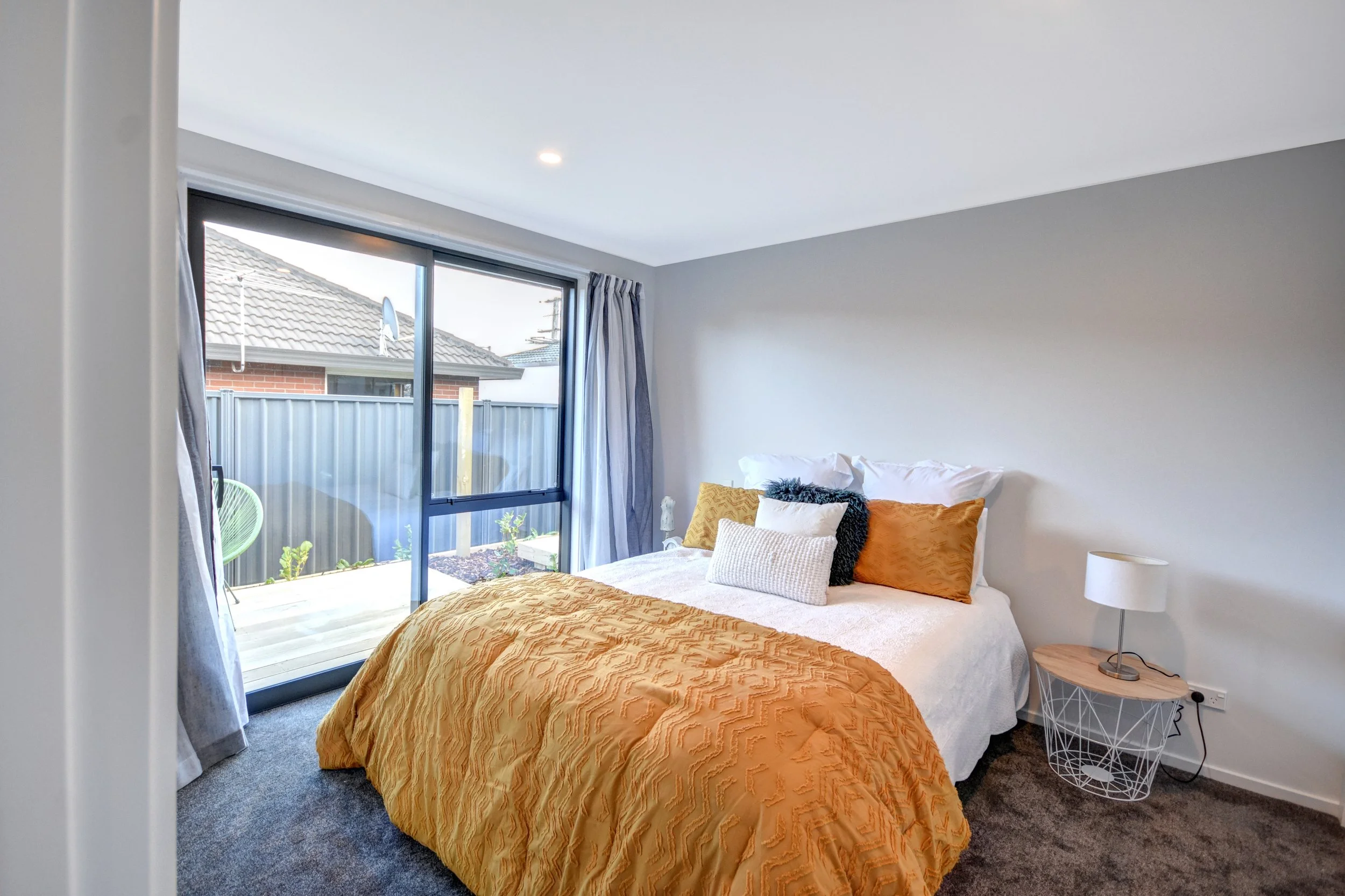 Bedroom with a bed covered in an orange textured blanket, multiple pillows, bedside table with a lamp, and a large sliding glass door leading to a small patio with plants.