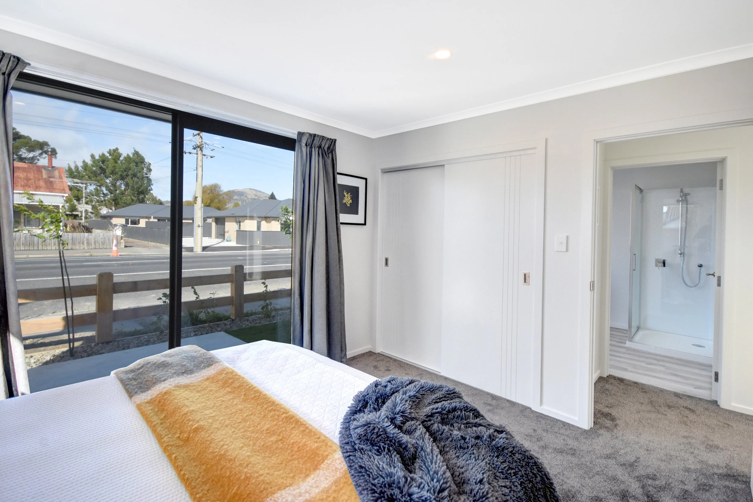 A bedroom with a large window showing a street view, a bed with a blanket, and an open door leading to a bathroom with a shower.