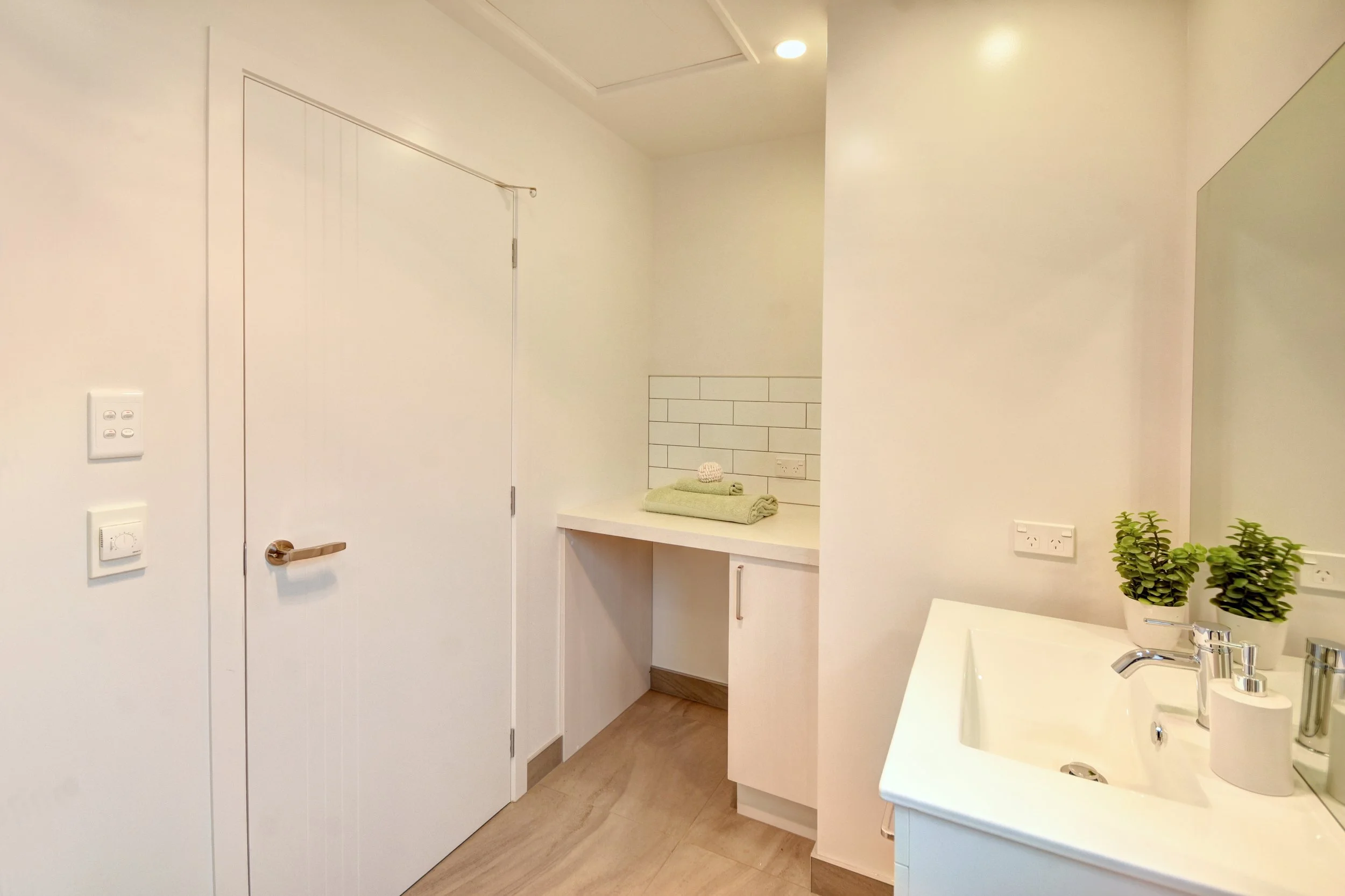 A clean, minimalist bathroom with white walls, a white door, a small countertop with folded towels, a white sink with soap dispensers, and potted green plants.