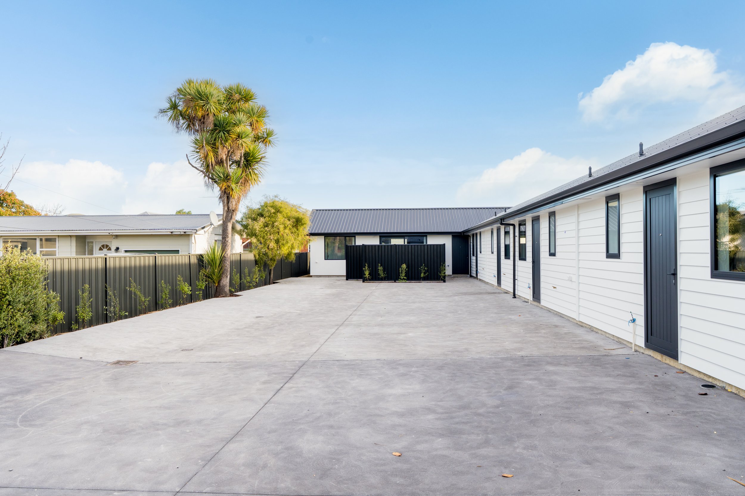 A backyard with a large concrete patio, white house with black window frames and doors on the right, black fence, and a tall palm tree on the left under a blue sky with some clouds.