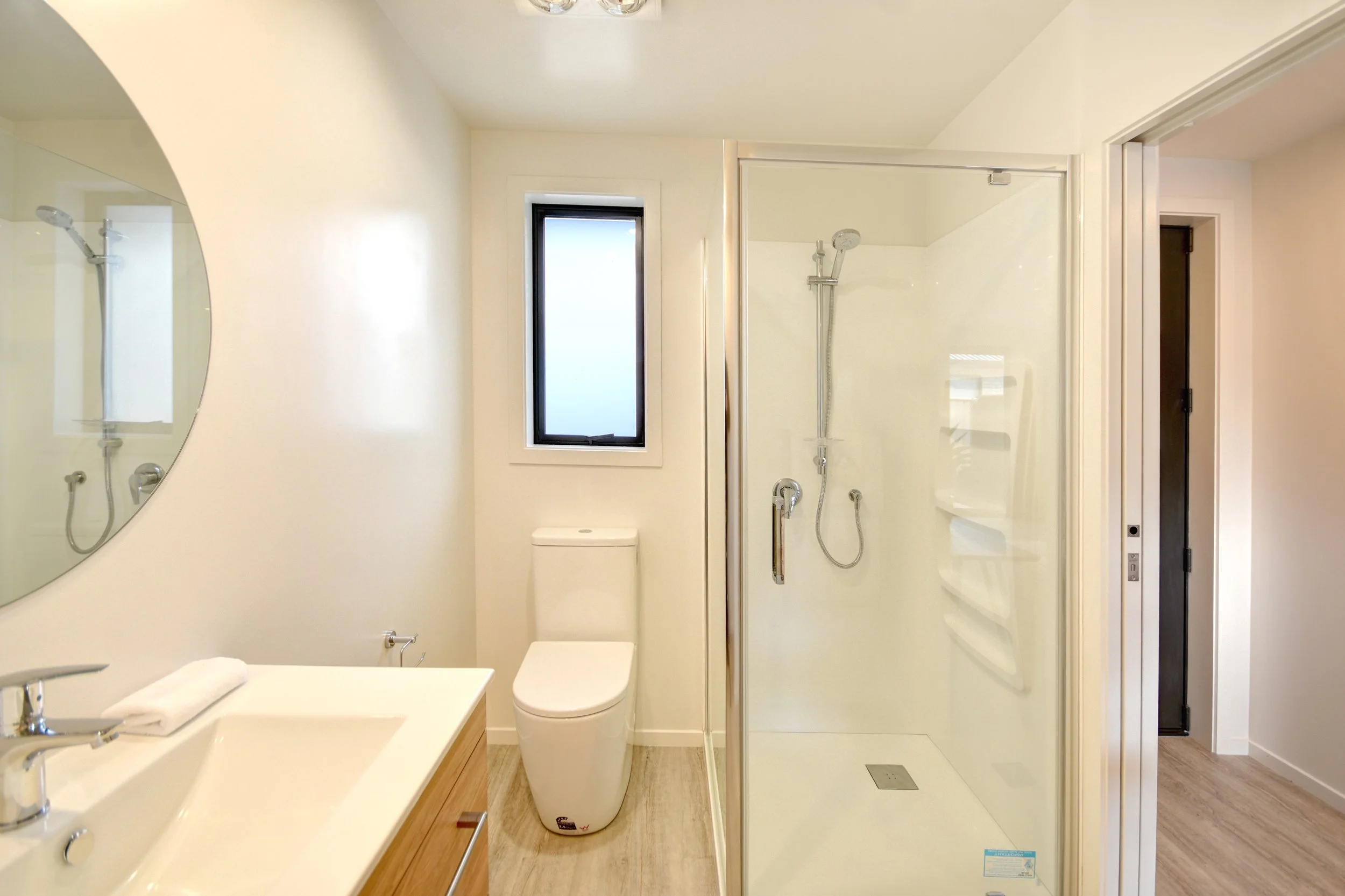 Modern bathroom with a mirror, sink, toilet, and shower with a glass door, small window, and wood-like flooring.