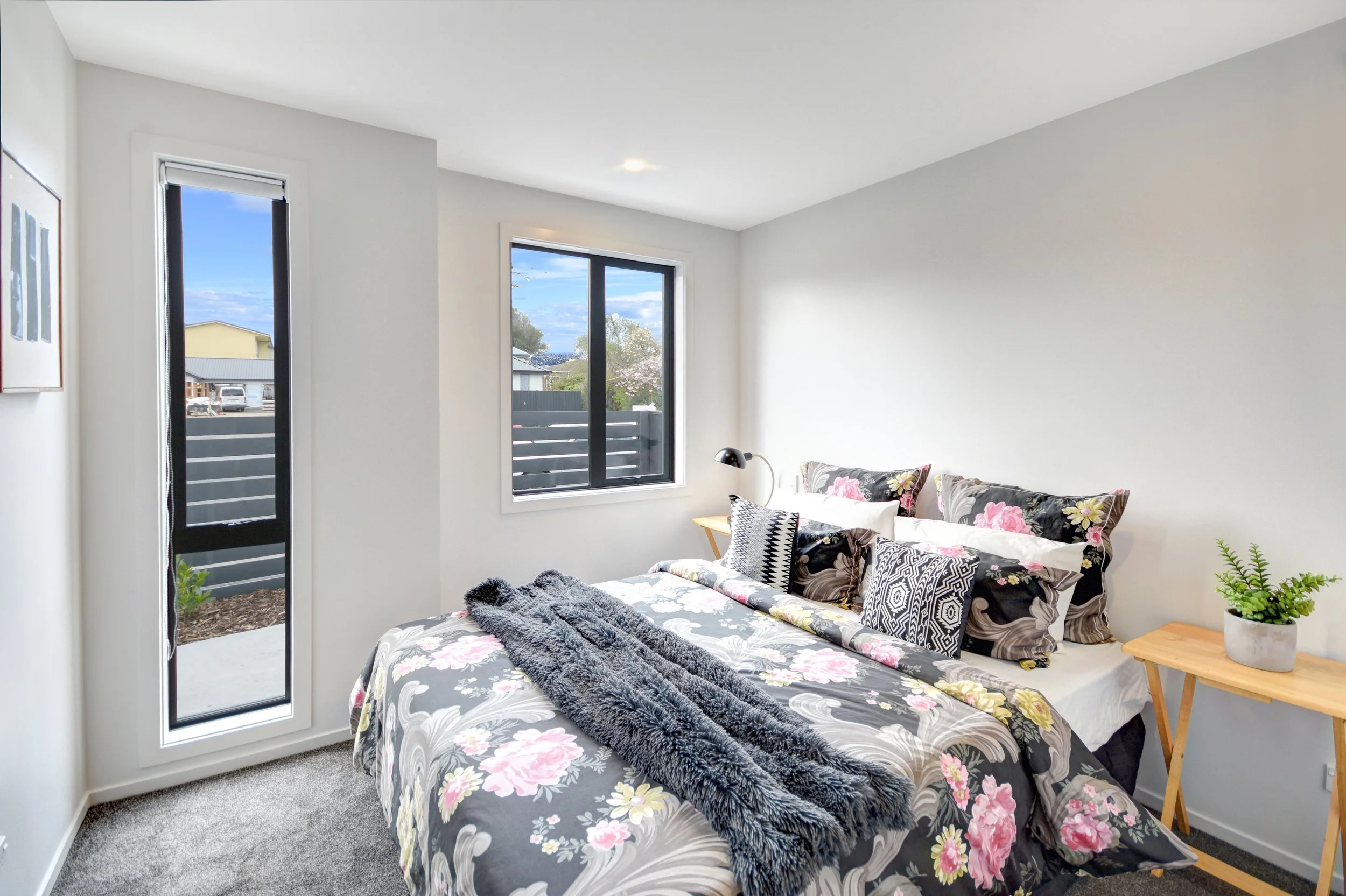 Bedroom with white walls, large windows, a bed with floral bedding and assorted pillows, a wooden nightstand with a small potted plant, and a small wooden side table with a lamp.