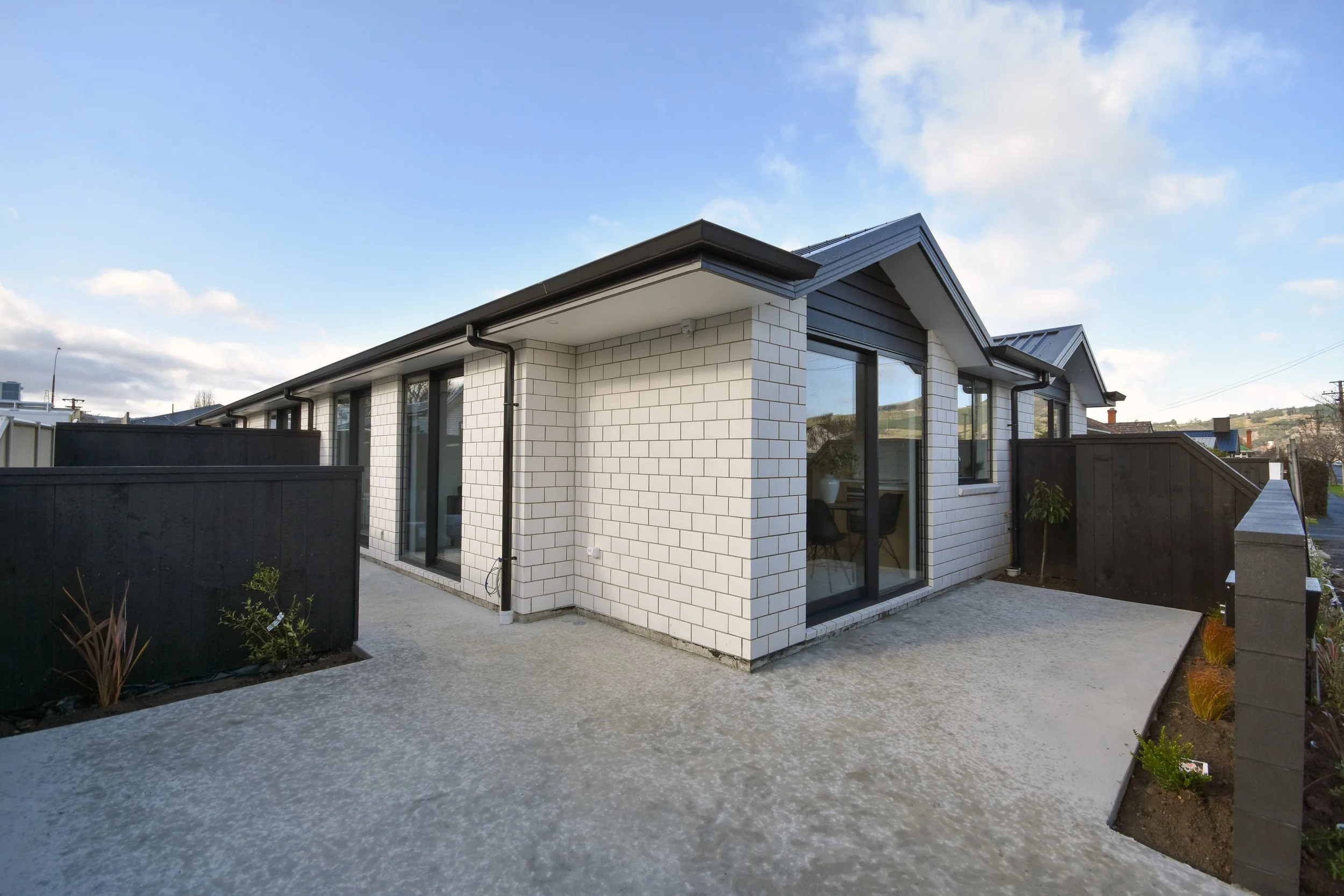 Modern single-story house with white brick walls, black roof, large glass sliding doors, and a small outdoor patio area, with a clear sky and some greenery visible.