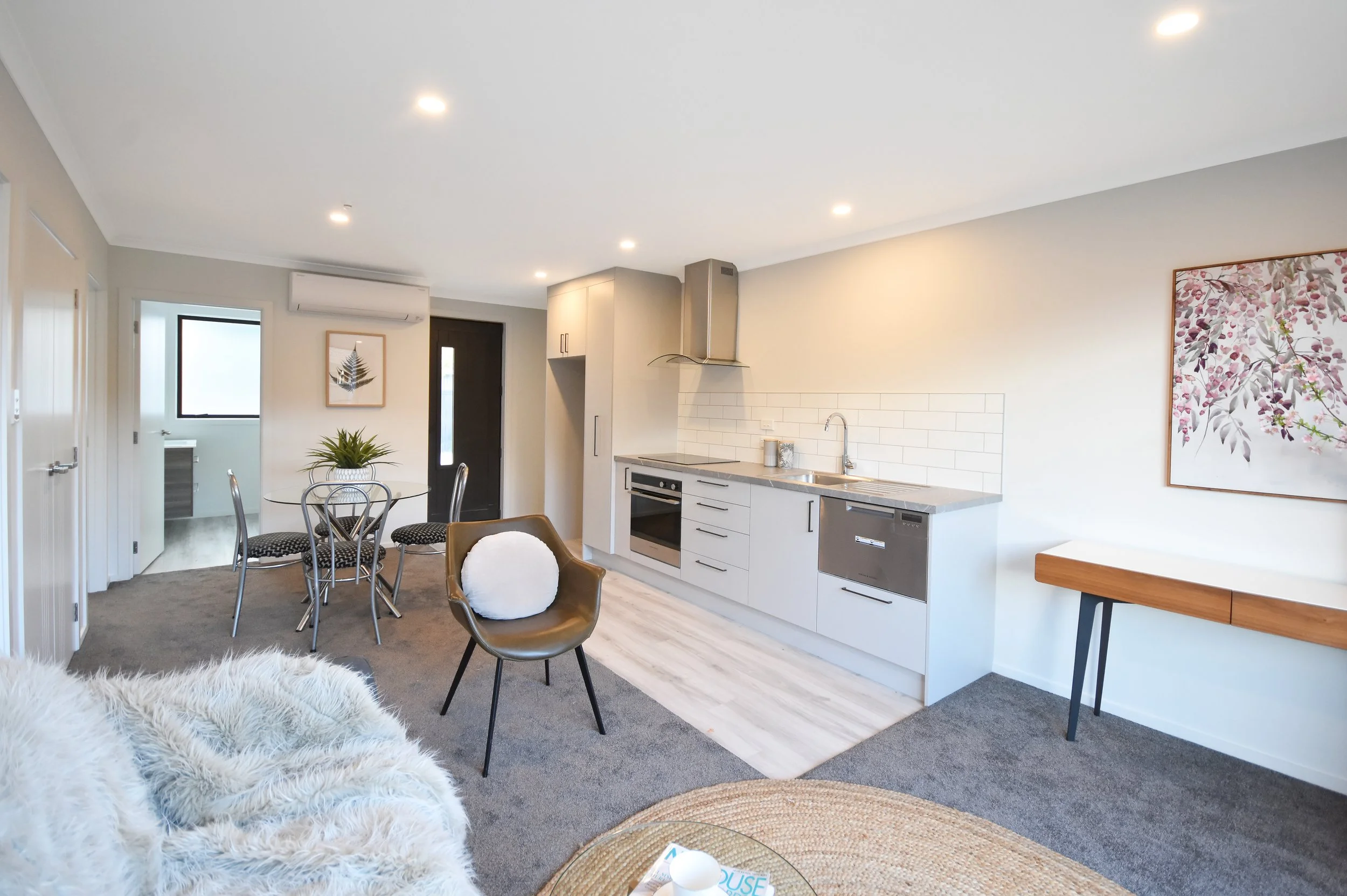 Open-concept living room and kitchen with white cabinetry, gray countertops, a stainless steel oven, a black front door, a round dining table with four chairs, a tan chair with a white cushion, a cozy fur blanket on a sofa, a glass-topped coffee tabl