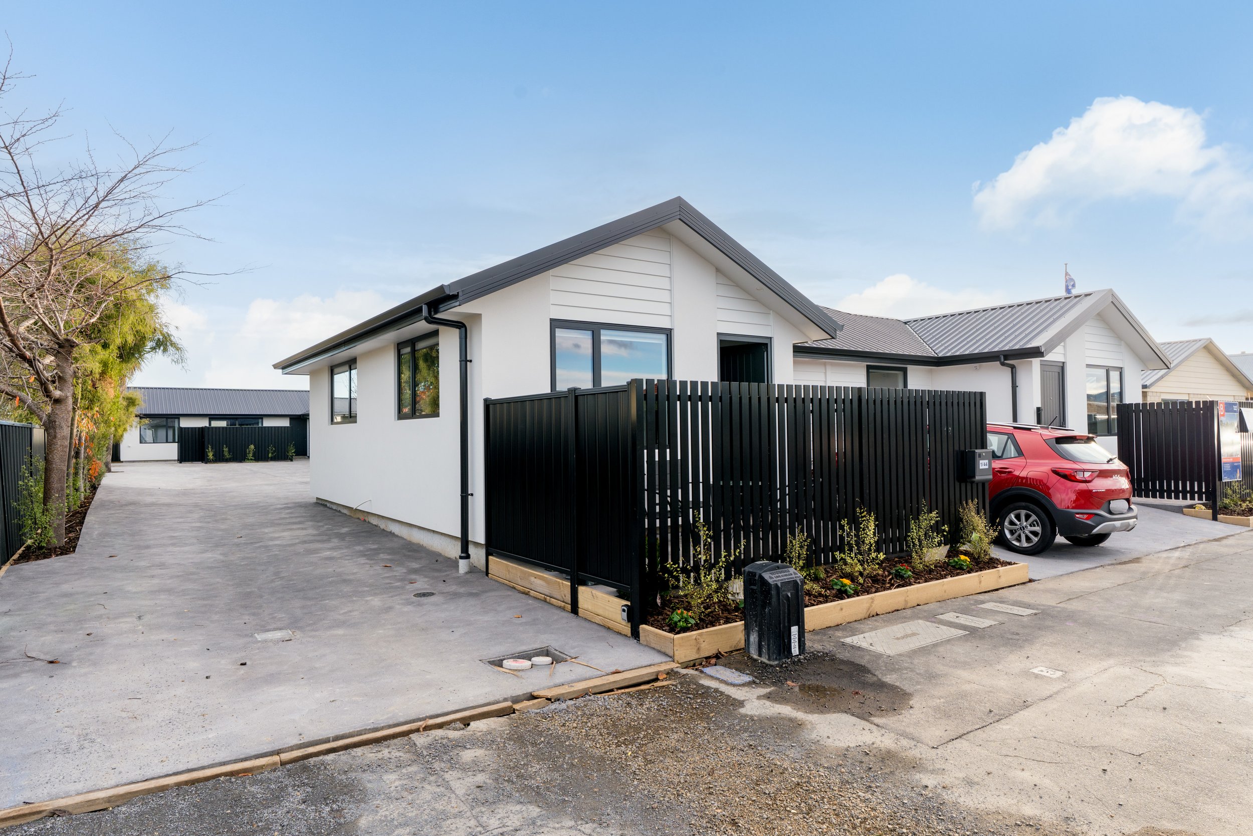 Modern white house with black roof, black fence, and a red SUV parked outside, with a landscaped garden and a clear blue sky.