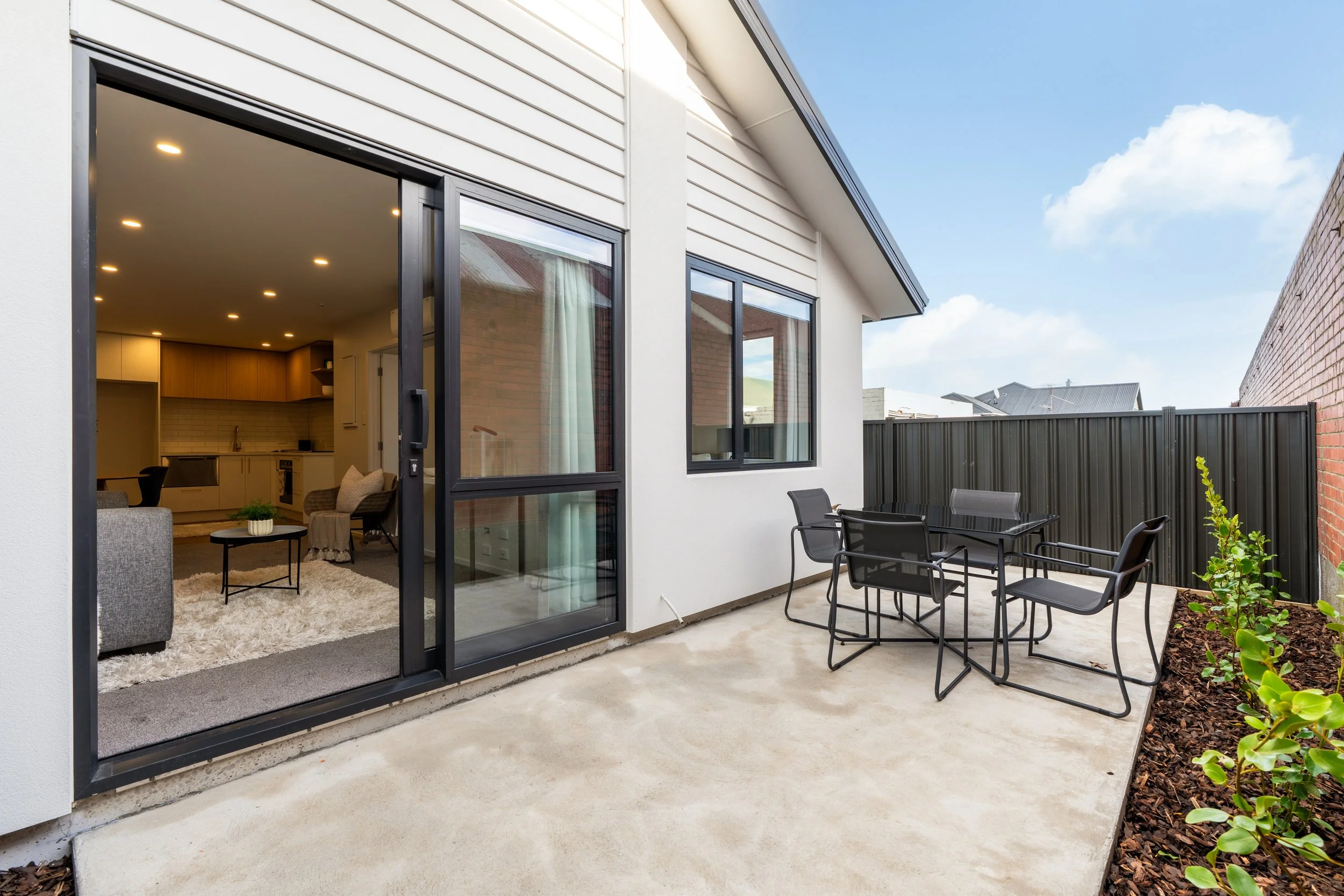 Outdoor patio with black metal table and chairs, fenced backyard, sliding glass door leading to a living room, plants along the edge of the patio, blue sky with clouds.