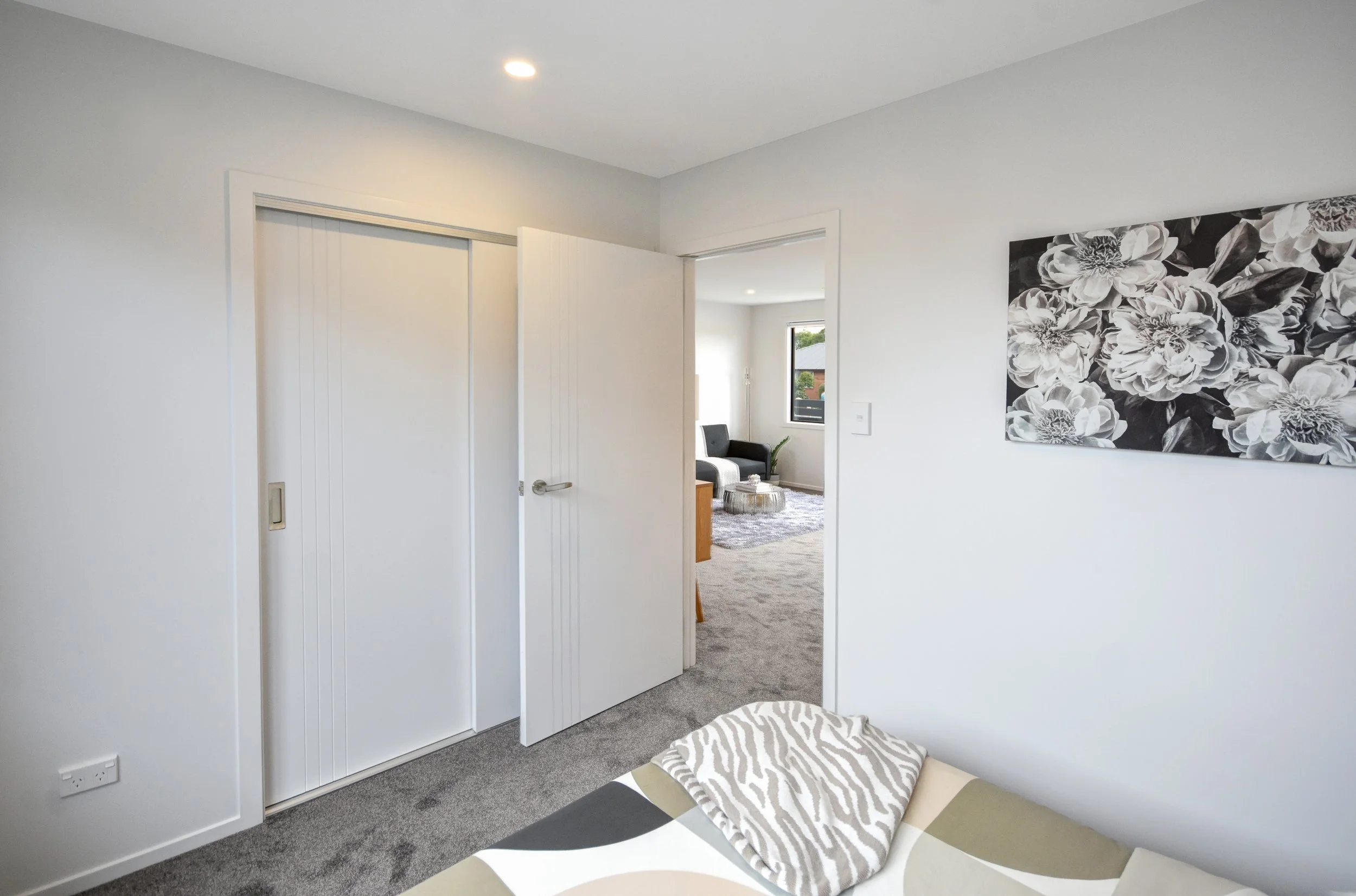 A bedroom with white walls, gray carpet, a bed with a blanket, and a black and white flower wall art. An open door reveals a living room with a black sofa and window.