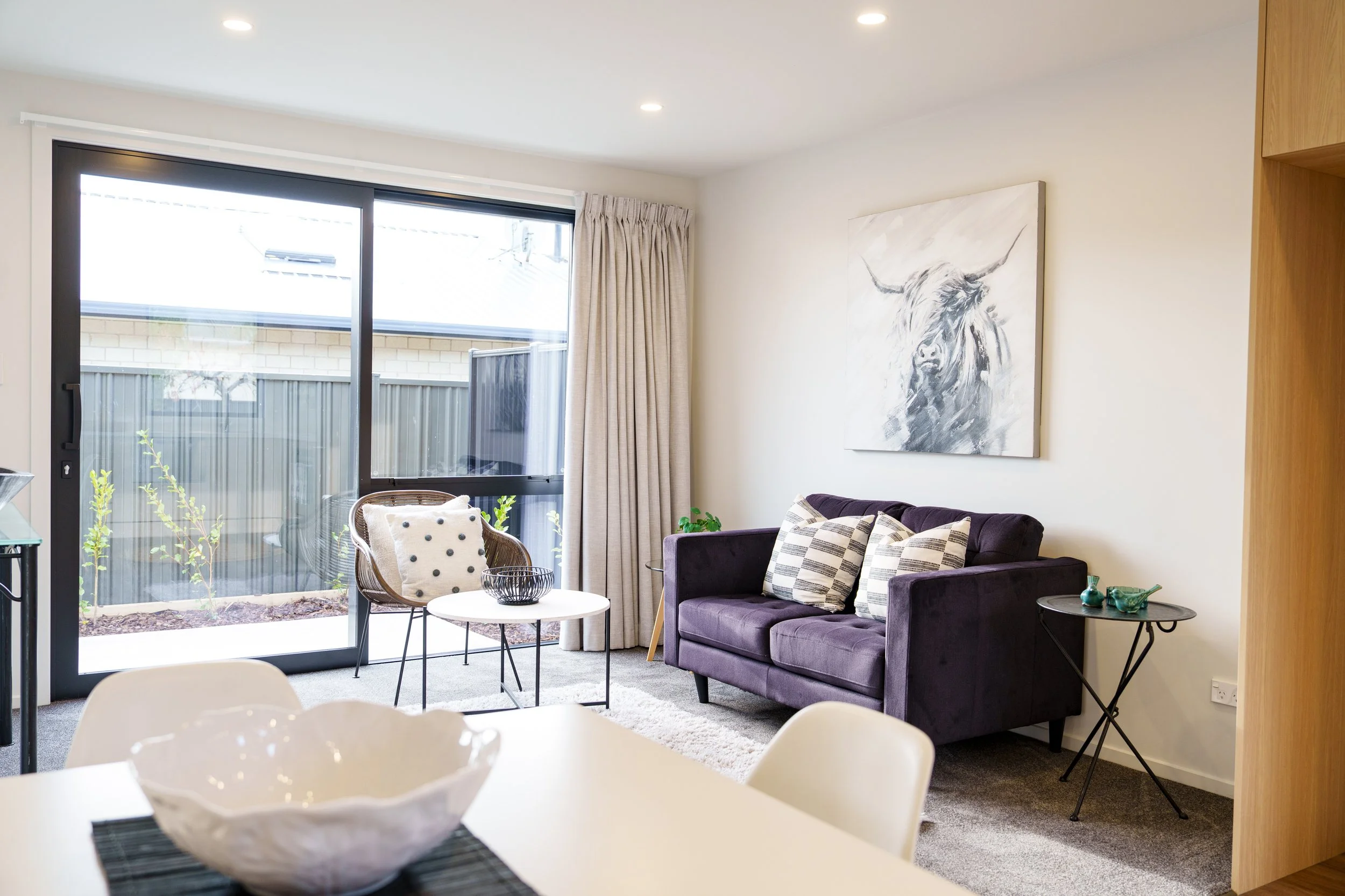 Living room with purple sofa, white wall art, glass door with curtains, small side tables, and decor items.