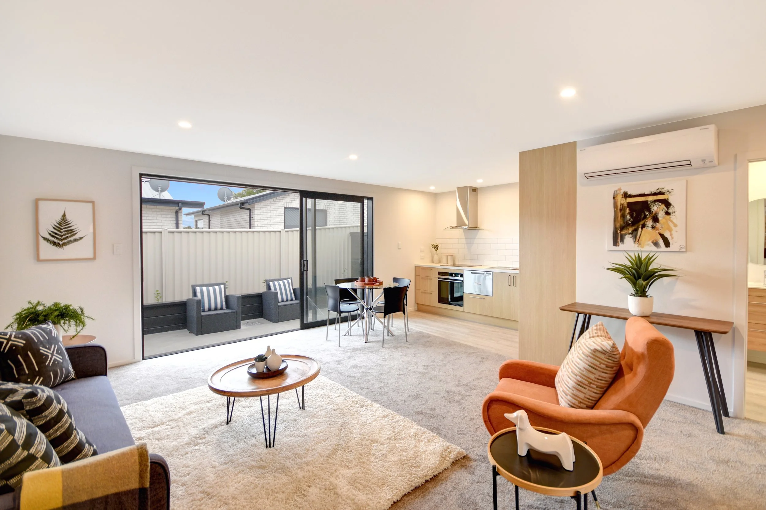Spacious living room with beige carpet, light wood furniture, orange and black chairs, abstract wall art, potted plant, and sliding glass door leading to outdoor patio with two chairs.