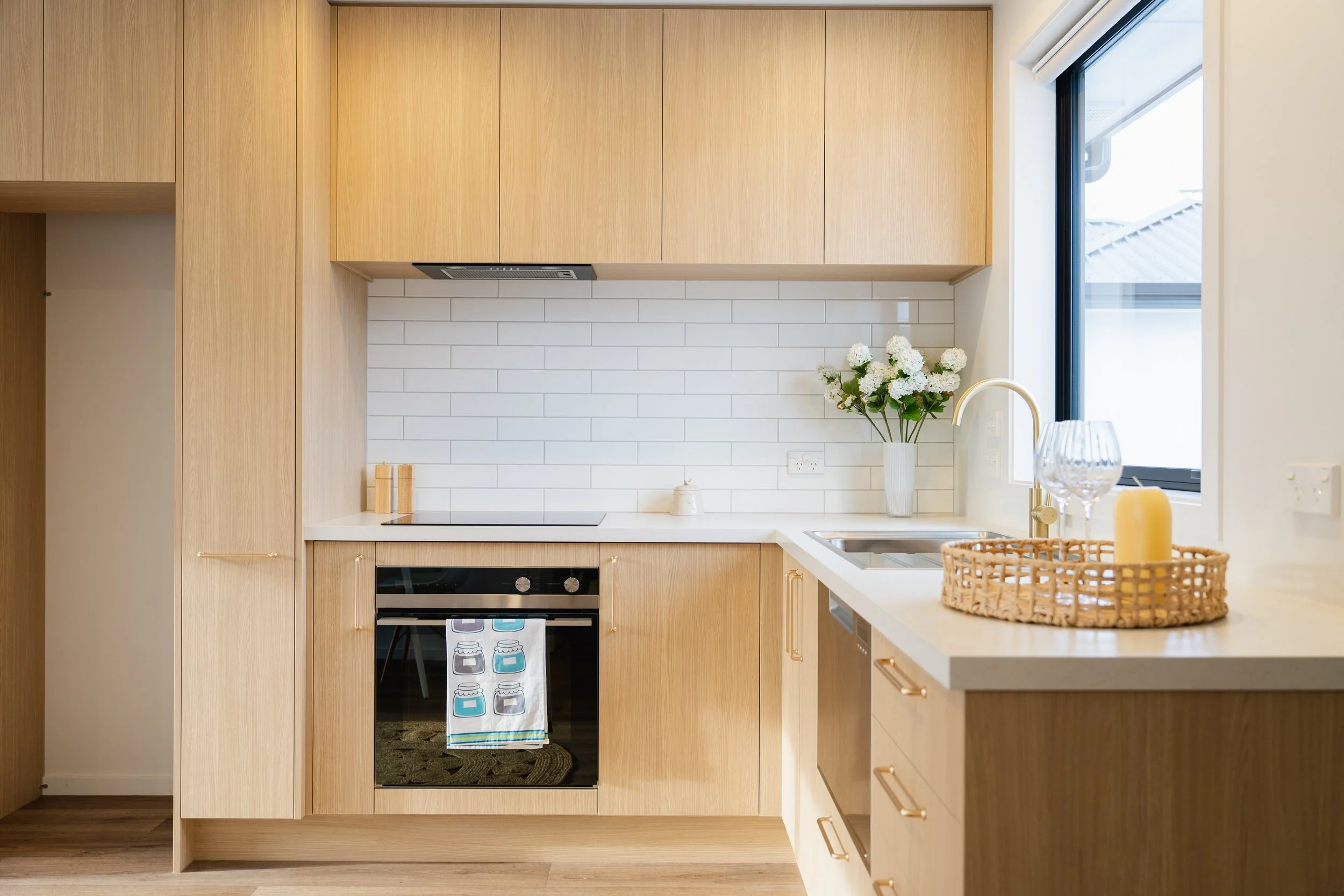Modern kitchen with light wooden cabinets, white backsplash, and a window. Decor includes a vase with white flowers, candles, and a tray with glasses and a candle.