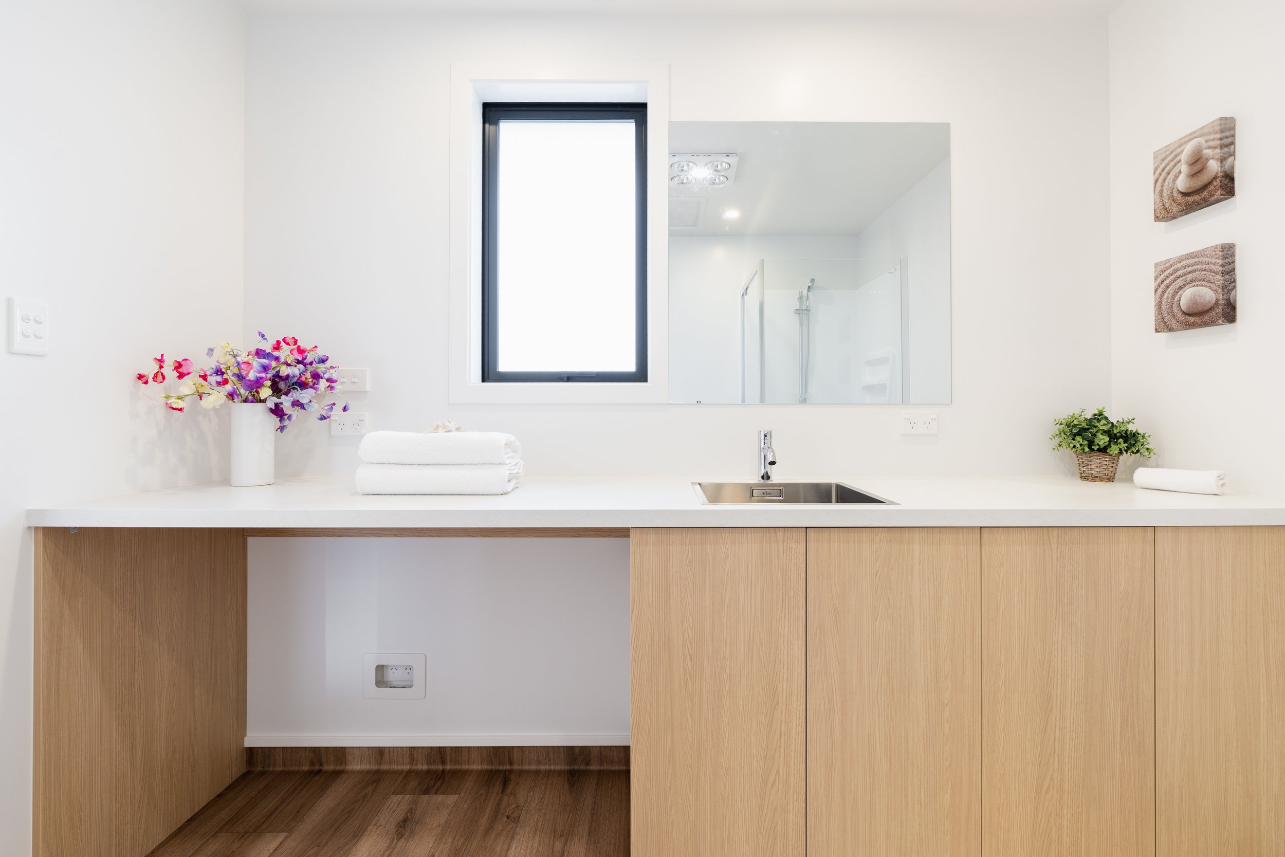 A minimalist bathroom vanity with a white countertop, wooden cabinets, a small sink, and a chrome faucet. Decor includes stacked white towels, a small basket of greenery, and a vase with purple and pink flowers. There is a window and a large mirror a