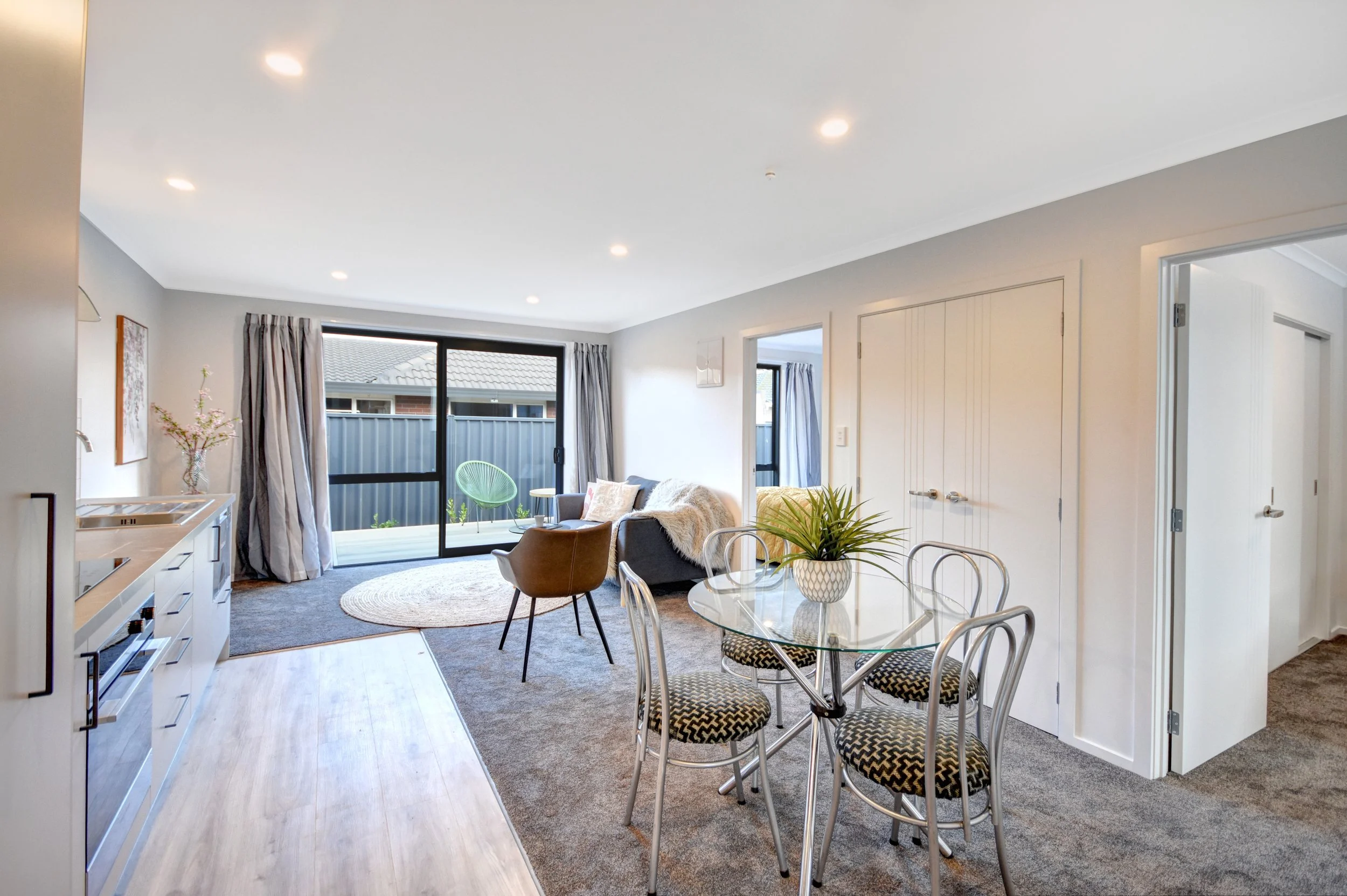 Living room and dining area with glass door opening to outdoor patio, modern furniture, potted plant, and kitchen area.