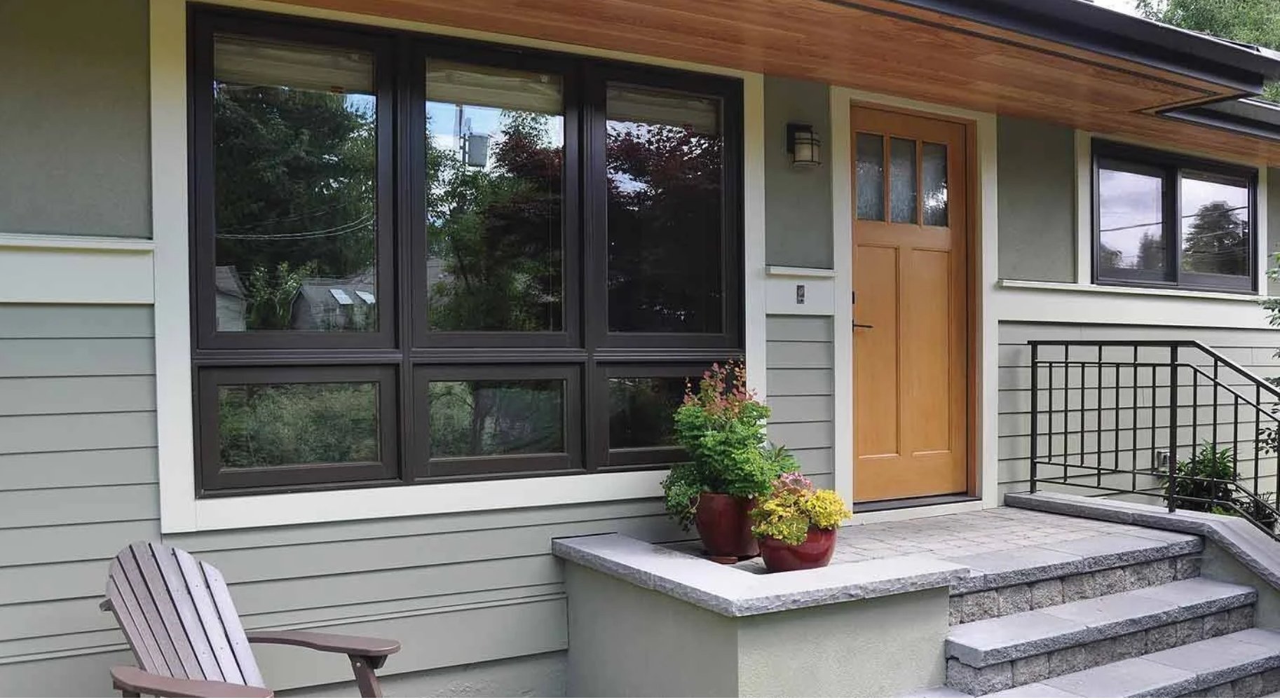 Front porch of a house with a wooden door, large window, potted plants, and a chair.