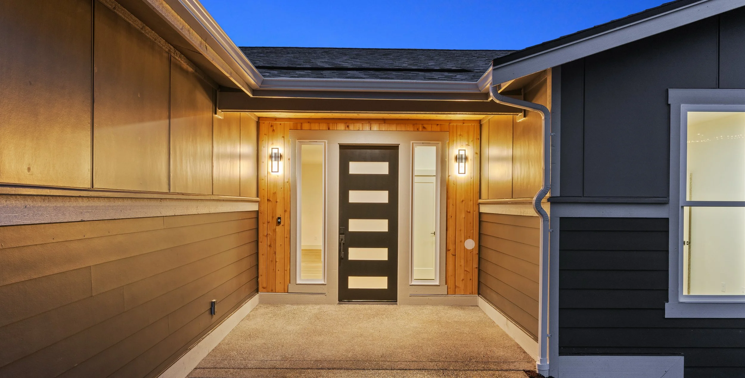 Modern front door with vertical rectangular glass panels, flanked by sidelights, on a house with wood and dark siding, illuminated by wall lights at dusk.