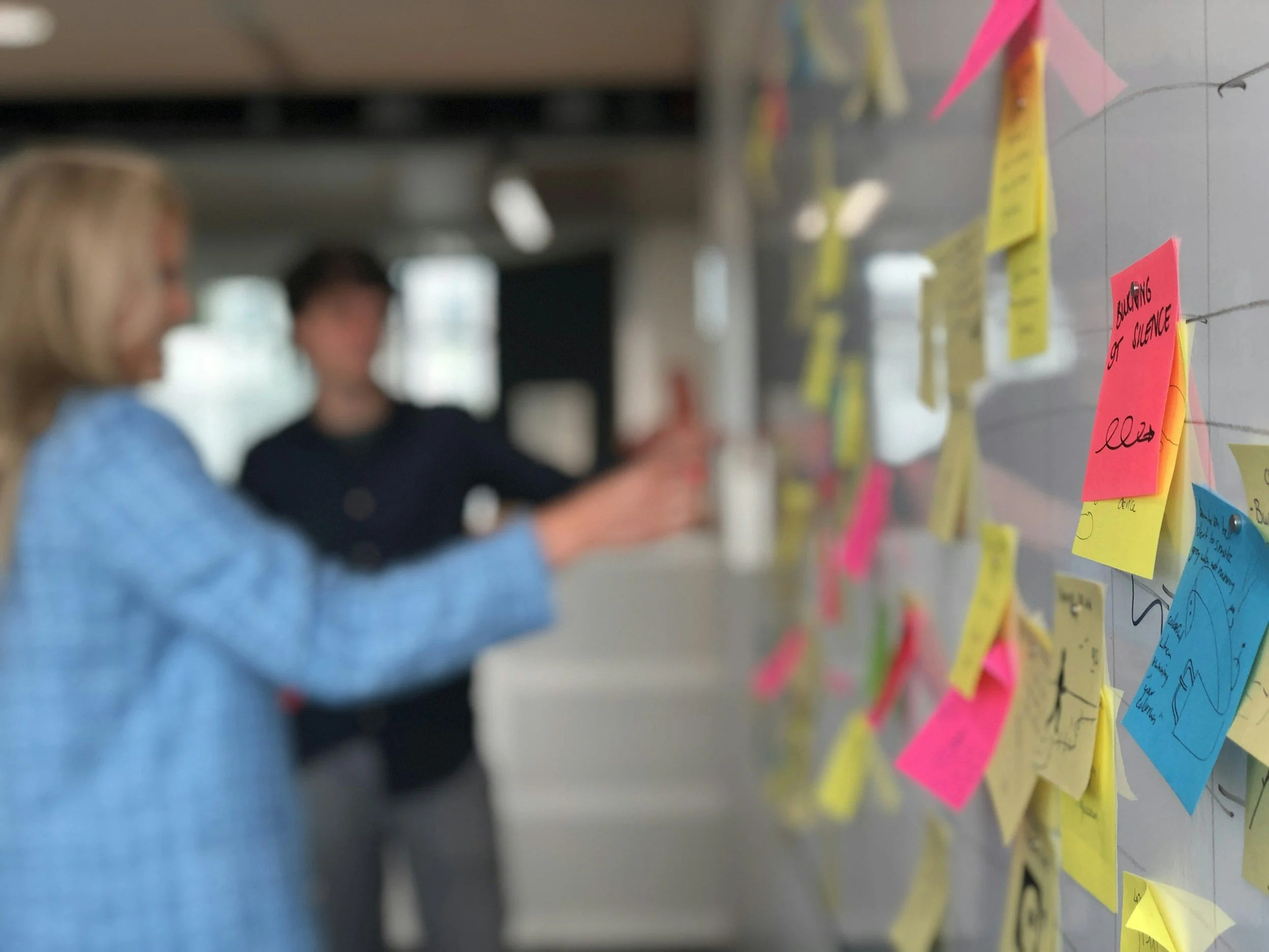 Close-up side view of a whiteboard with colored handwritten sticky notes being arranged by two humans, who's figures have been blurred for privacy.